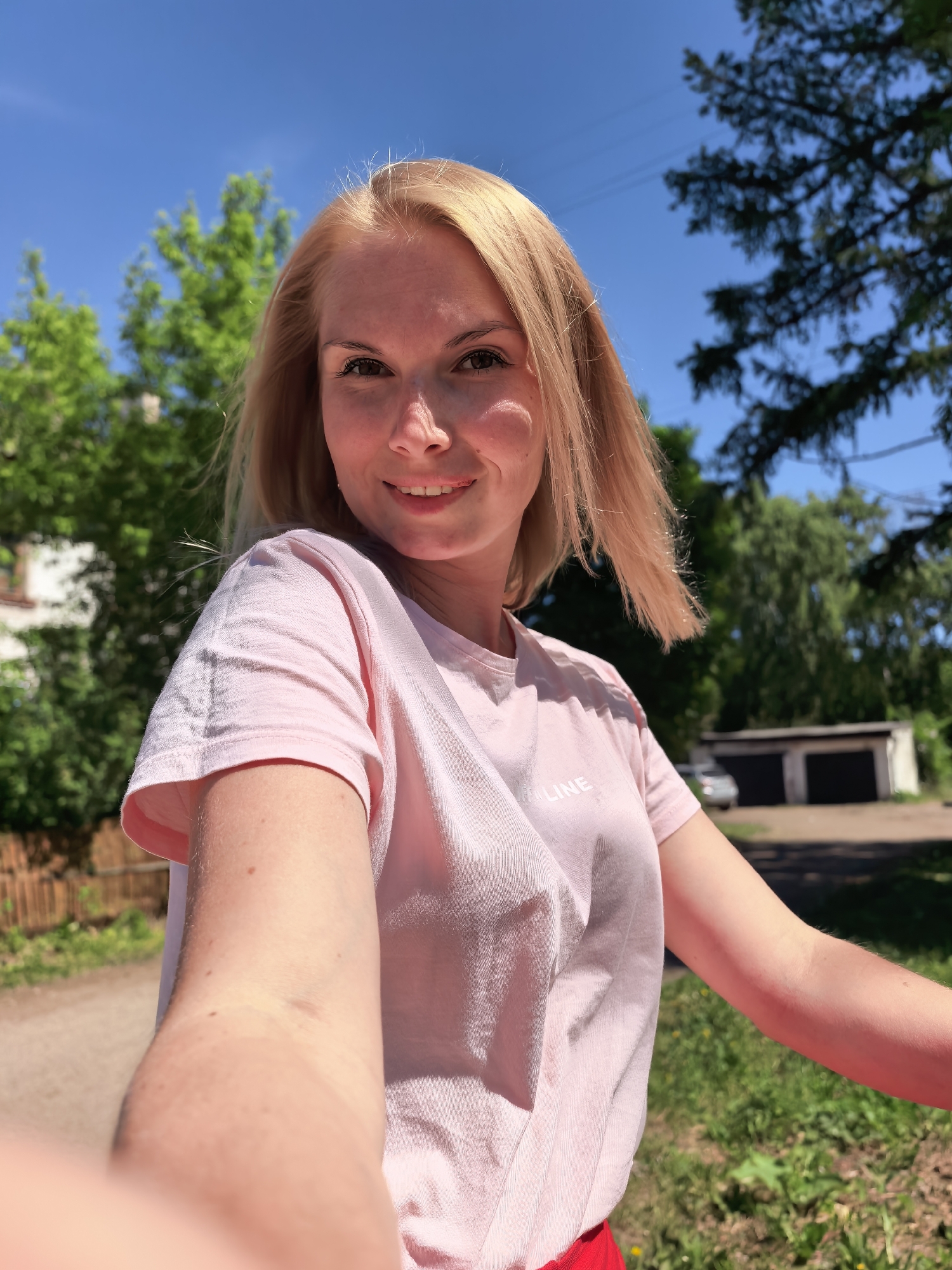 A fair-skinned woman with blonde, shoulder-length hair smiles directly at the camera. She is wearing a pink t-shirt and red bottoms. The photo appears to be a selfie taken outdoors in daylight. Behind her is a background of green trees, blue sky, a residential building, and a garage. The scene is potentially located in Sterlitamak, Russia.