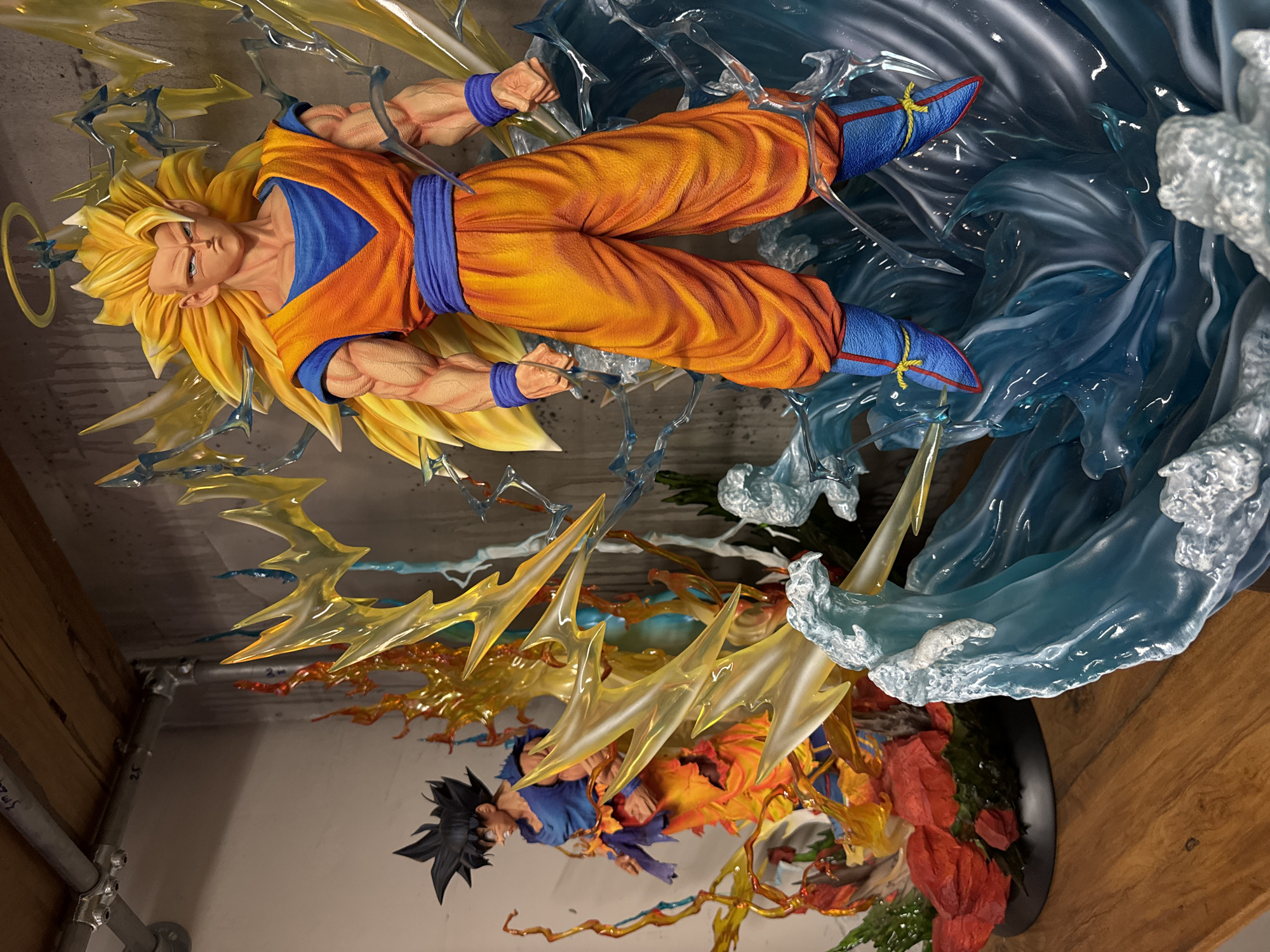 The image displays two large, detailed collectible figures from the Dragon Ball Z franchise, meticulously arranged on a wooden surface within an indoor setting in Đà Nẵng, Vietnam.
The foreground features a "Super Saiyan 3 Goku" figure, standing prominently. This character exhibits long, spiky golden-yellow hair, a stern facial expression with no eyebrows, and a golden halo above its head. It wears an orange gi, blue undershirt, blue waist sash, blue wristbands, and blue boots with red trim and yellow laces. The figure is positioned amidst translucent blue sculpted waves, representing water or energy, and is surrounded by sharp, translucent yellow and light blue energy effects suggesting intense power.
In the background, partially obscured, is another "Goku" figure, characterized by spiky black hair and a blue undershirt under a torn orange gi, with blue wristbands. This figure stands on a circular black base adorned with red sculpted elements resembling rocks or debris, and is engulfed by translucent orange and yellow flame-like energy effects, accented with subtle blue elements. Both figures are static displays, capturing dynamic poses of combat or power activation.
The environment consists of a polished medium-brown wooden surface serving as a display base. The background wall is light grey, likely exposed concrete, showing minor texture and vertical streaks. Above the figures, an industrial-style overhead structure is visible, composed of galvanized steel pipes and fittings supporting a raw wooden shelf. Handwritten markings, including "sm24" and "25," are visible on the pipes and shelf. The scene is well-lit, emphasizing the figures' details and translucent effects. This setting suggests a dedicated display area, likely within a modern retail establishment specializing in collectibles, an anime merchandise store, or a private collection displayed in a loft-style or industrially designed room in an urban area of Đà Nẵng.