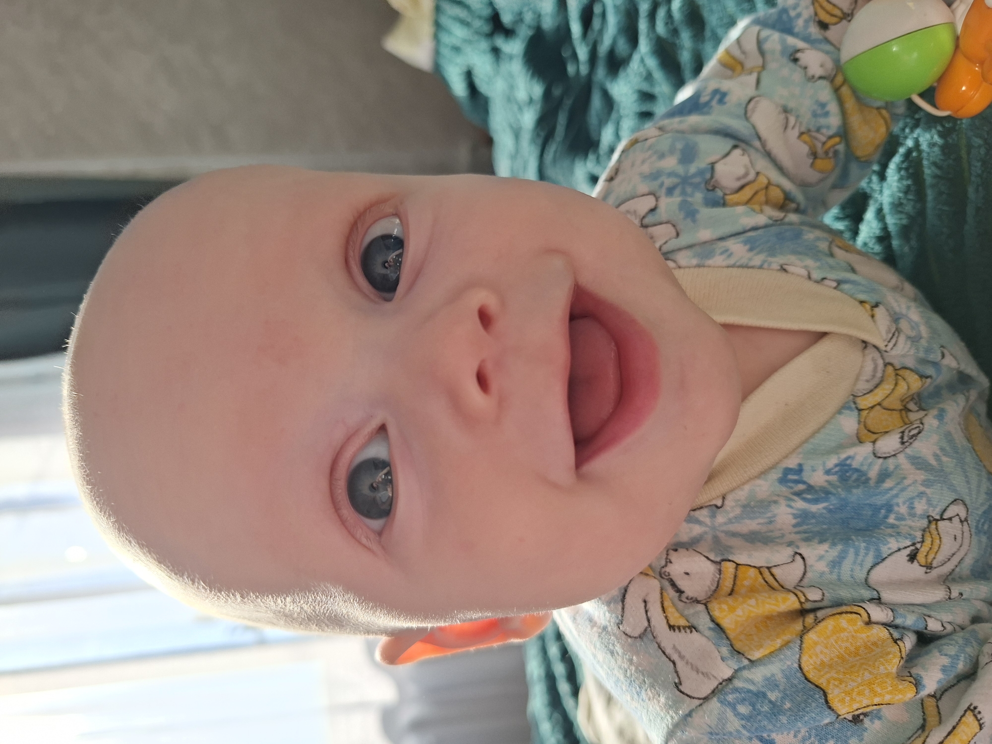 A baby lies on a surface, wearing a light blue patterned onesie with white and yellow bear designs. The baby has pale skin, a bald head with fine blonde hair, and blue eyes. The baby's mouth is open. A colorful toy is visible in the upper right corner. The background includes a teal, textured fabric. The location is Unknown, Unknown.