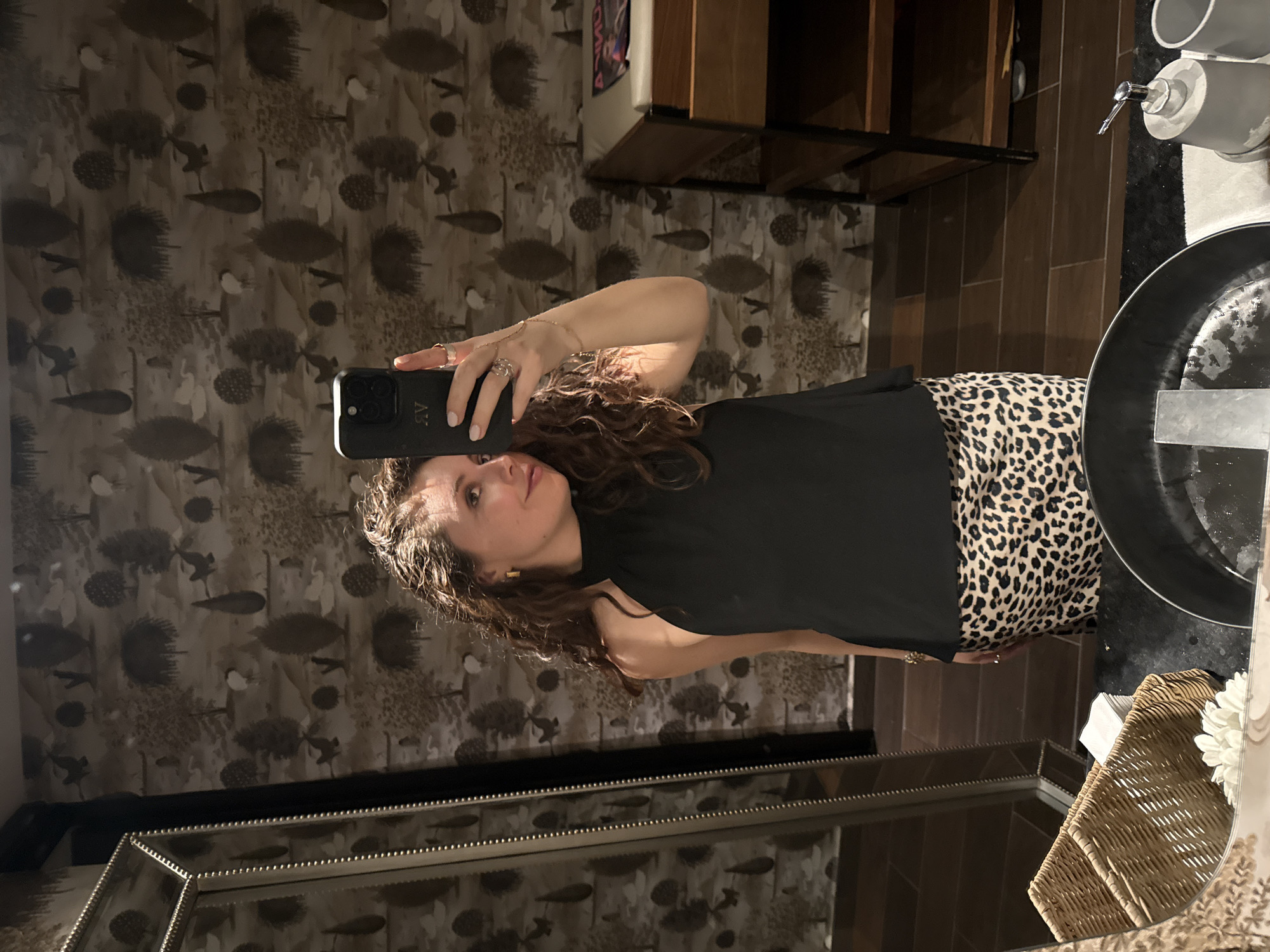 A woman with curly hair is taking a selfie in a bathroom, likely in a home. She is wearing a black top and a leopard-print skirt. The background shows a mirror with a decorative frame, wallpaper with a nature-inspired pattern (trees and birds), a sink with a black countertop, a soap dispenser, and a small wooden shelf unit. A wicker basket with a white flower is partially visible on the right side of the mirror's reflection. The photograph was probably taken in Puebla, Mexico, as implied by the prompt.