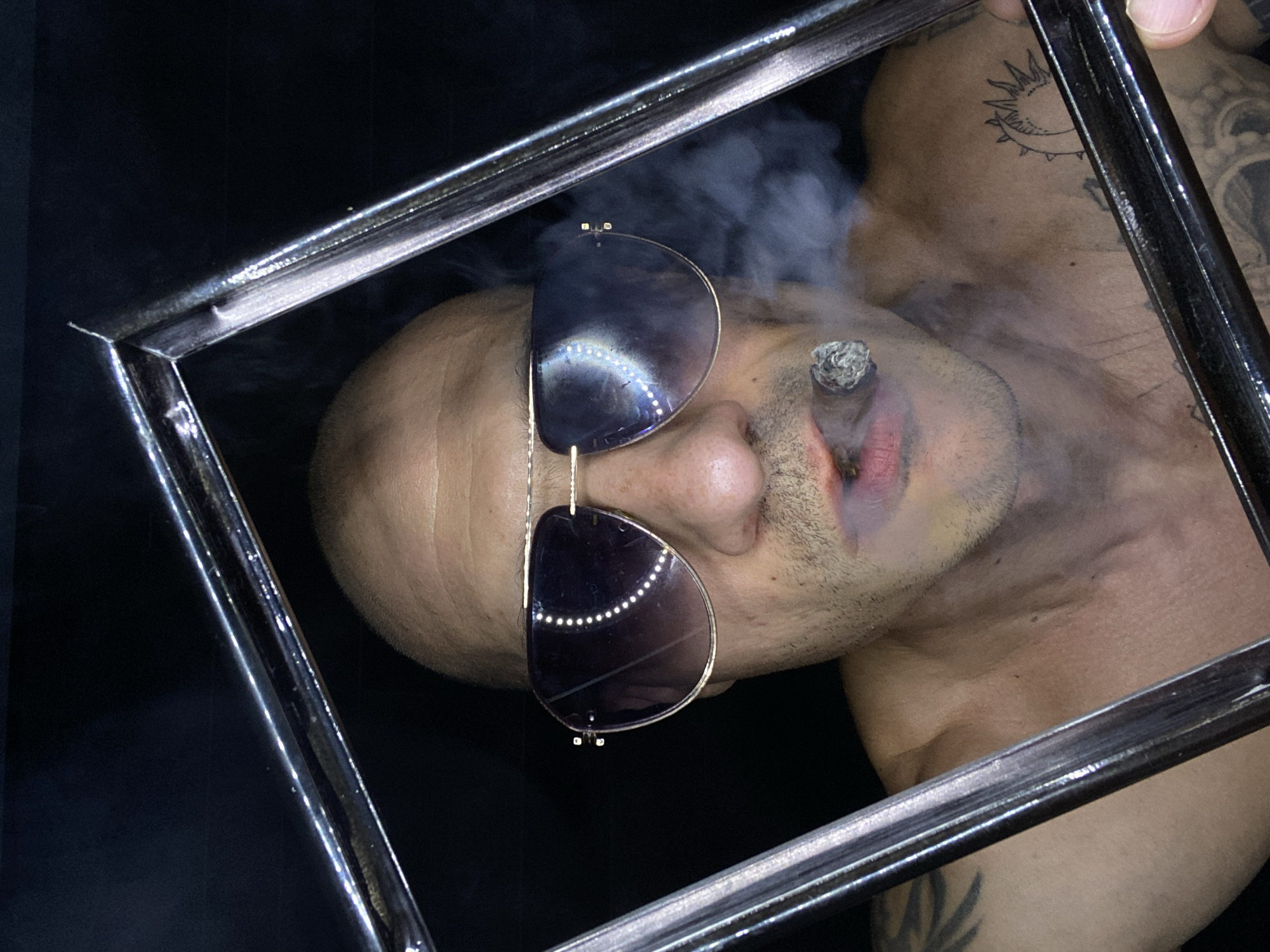 A shirtless, bald man with tattoos and sunglasses is shown through a silver square frame, smoking a cigar. The background is black. The exact location is not discernible, but it is stated to be in the city of Pleven, Bulgaria.