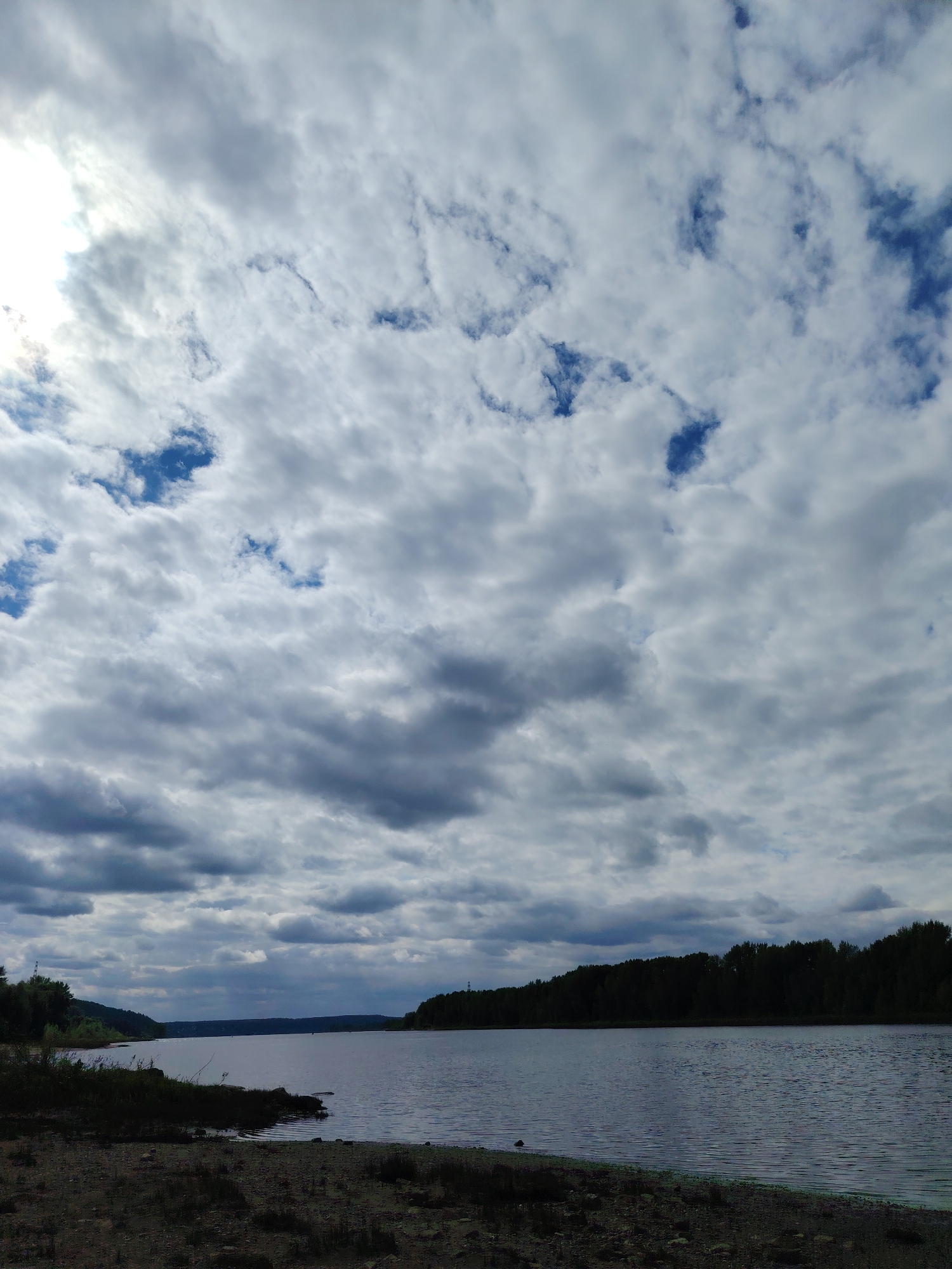 This is a vertical, full-shot, eye-level photograph of the Kama River embankment in Chaikovsky, Russia, under a cloudy sky. The foreground features a rocky and sandy shore. The middle ground is dominated by the wide, still surface of the Kama River. In the background, a dense line of dark green trees forms a treeline along the opposite bank of the river, suggesting a rural or natural landscape. The sky is filled with a dramatic display of white and gray clouds, with patches of blue sky visible in the upper portion. The lighting is diffused due to the cloud cover, casting a subdued light over the scene. There are no people or animals visible in the image. The overall mood is serene and natural.