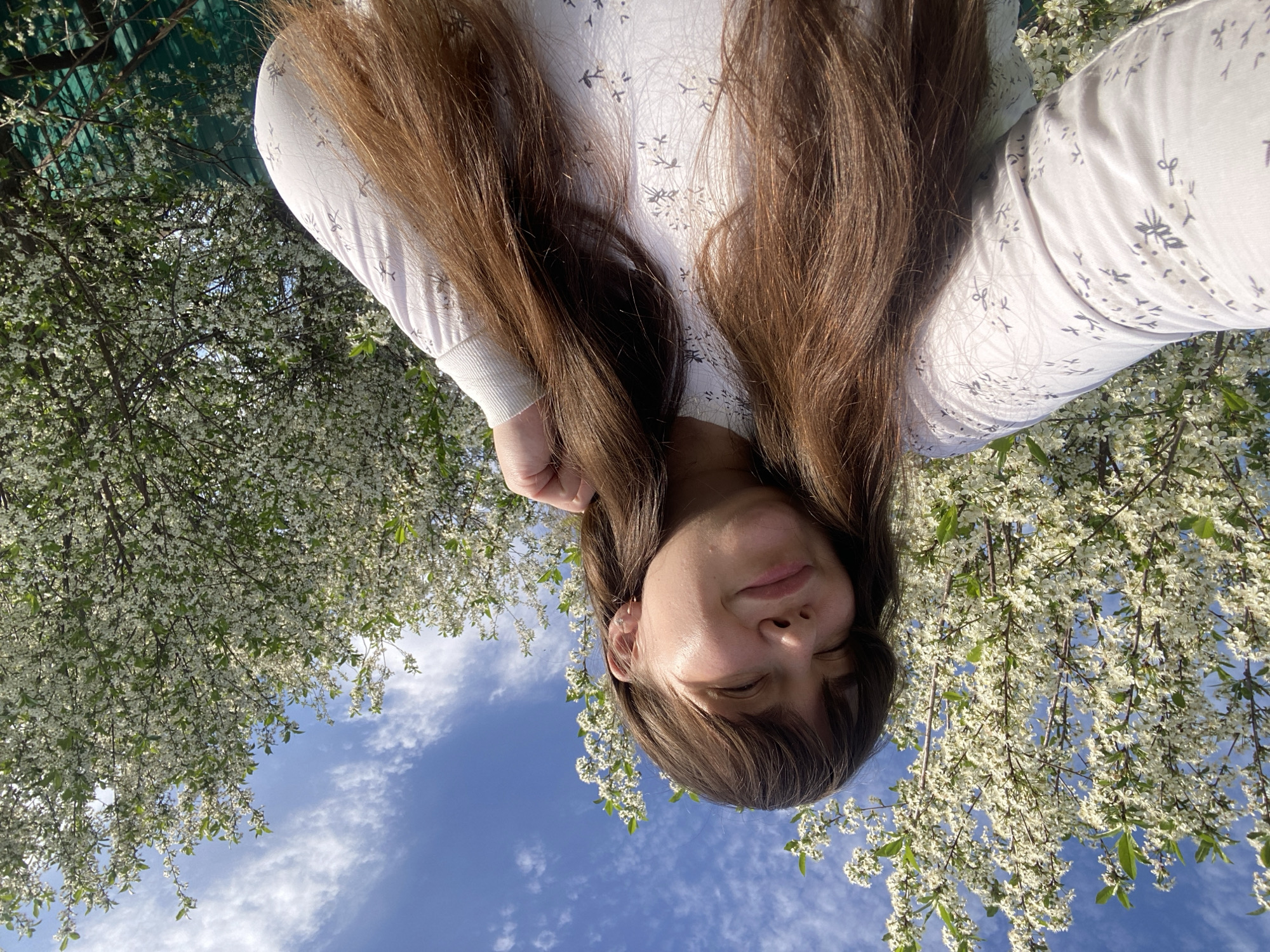A woman with long brown hair is positioned upside down in the foreground. She is wearing a long-sleeved white shirt with a small, repeating pattern. Her eyes are mostly closed.
The background consists of a blue sky with scattered clouds and blooming trees with white flowers. Some green netting is visible in the upper left corner.
Given the prompt about Votkinsk, Russia, the location is likely a park or garden in the city during springtime when trees are in bloom.