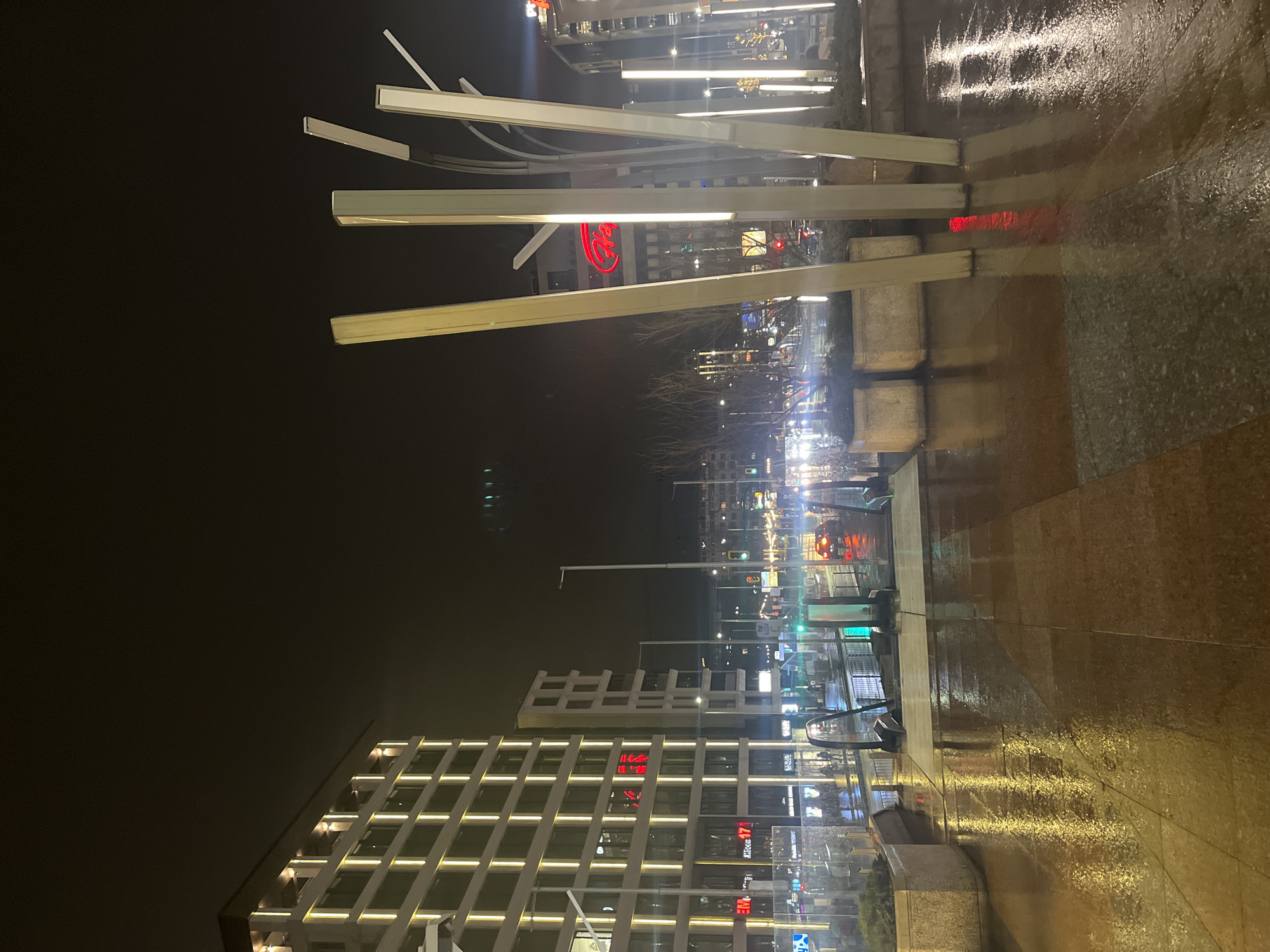 The image captures a nighttime scene in Sofia, Bulgaria, likely on a wet surface given the reflections. In the foreground, there are illuminated, modern art installations comprised of horizontal rectangular structures that light up the area and reflect on the wet pavement. Further back, there are modern buildings with distinctive lit-up grid-like facades. In the distance, city lights and traffic are visible. The wet surface amplifies the illumination, creating a vibrant and somewhat distorted reflection of the surrounding environment. No people are clearly identifiable, suggesting a low-traffic or after-hours setting.