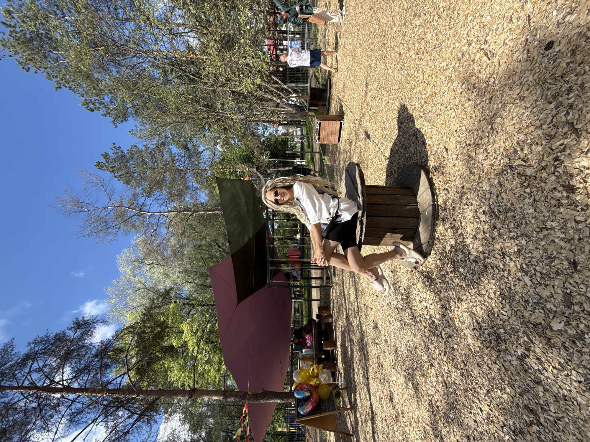 The image shows a woman with blonde dreadlocks sitting on a wooden cable reel in an outdoor setting. She is wearing sunglasses, a white top, and black shorts with white sandals. She is looking directly at the camera. The ground is covered in wood chips. In the background, there is a shaded area with tents and balloons, and other people are standing around. The trees and clear blue sky suggest a park or outdoor event. Based on the location information provided, this could be taking place in Rusanovo, Russia.