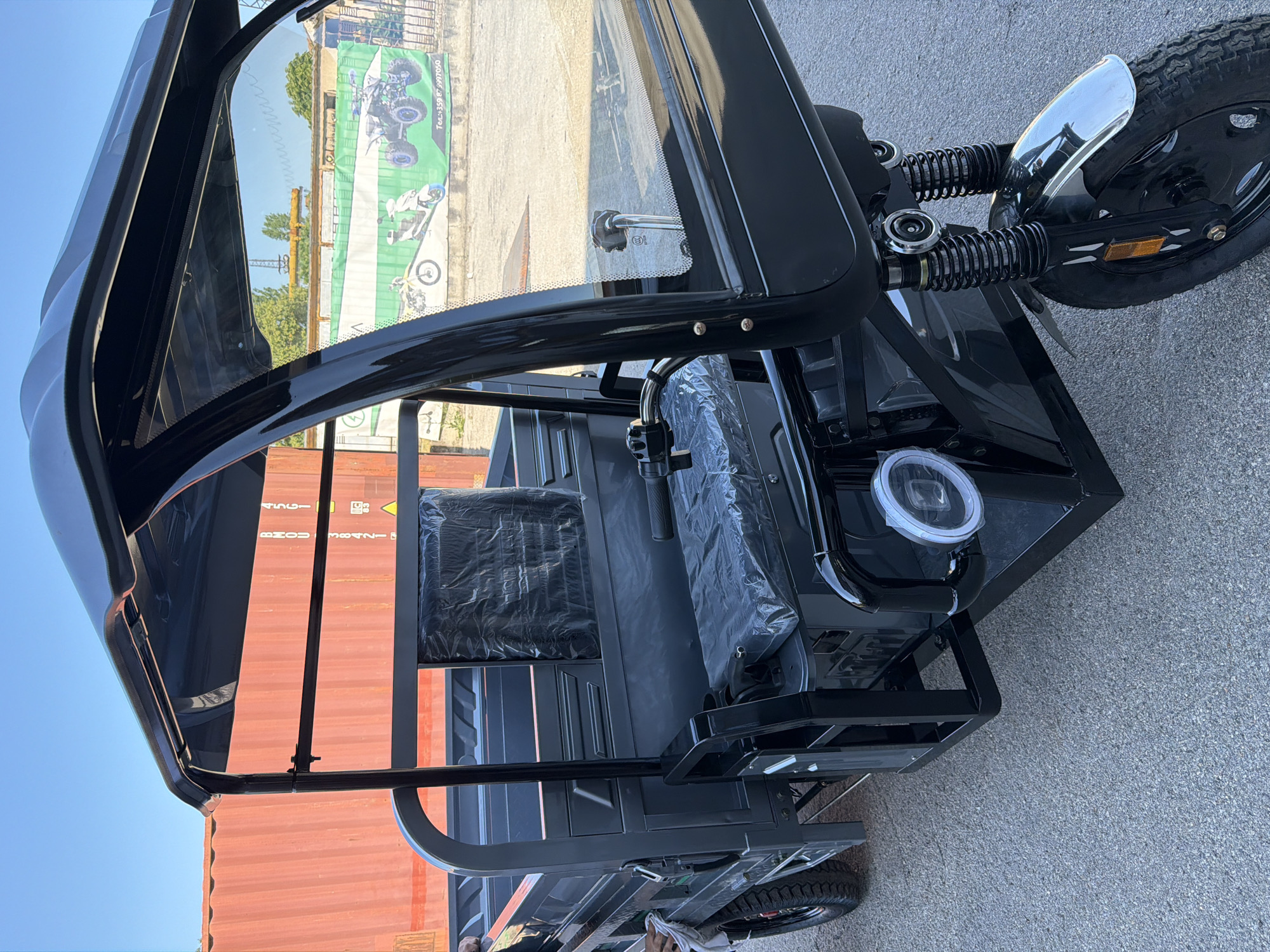 An outdoor, eye-level, medium shot captures the front right quarter of a black three-wheeled electric vehicle. The vehicle is parked on a gray, asphalt surface. The vehicle features a black canopy supported by a metal frame, and a black windshield. A black seat, covered in plastic, is visible behind the handlebars. A round, white headlight with a hexagonal lens is mounted on the front of the vehicle. The front wheel, with a chrome fender, is visible. In the background, an orange shipping container stands to the left, and a green banner with images of motorcycles and ATVs is displayed on a building to the right. The sky is clear and blue.
