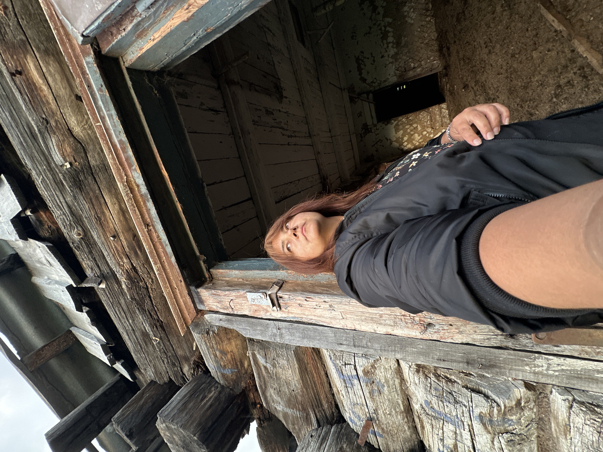 A woman with shoulder-length reddish-brown hair stands in the doorway of a dilapidated wooden structure, looking upwards and to the right. She is wearing a black bomber jacket over a floral patterned shirt and has on a silver bracelet on her left wrist. The structure appears to be made of rough-hewn logs and beams, with visible signs of decay and weathering. The doorway itself is partially open, revealing a dark interior with a rough floor. The overall impression is one of an abandoned or derelict building.