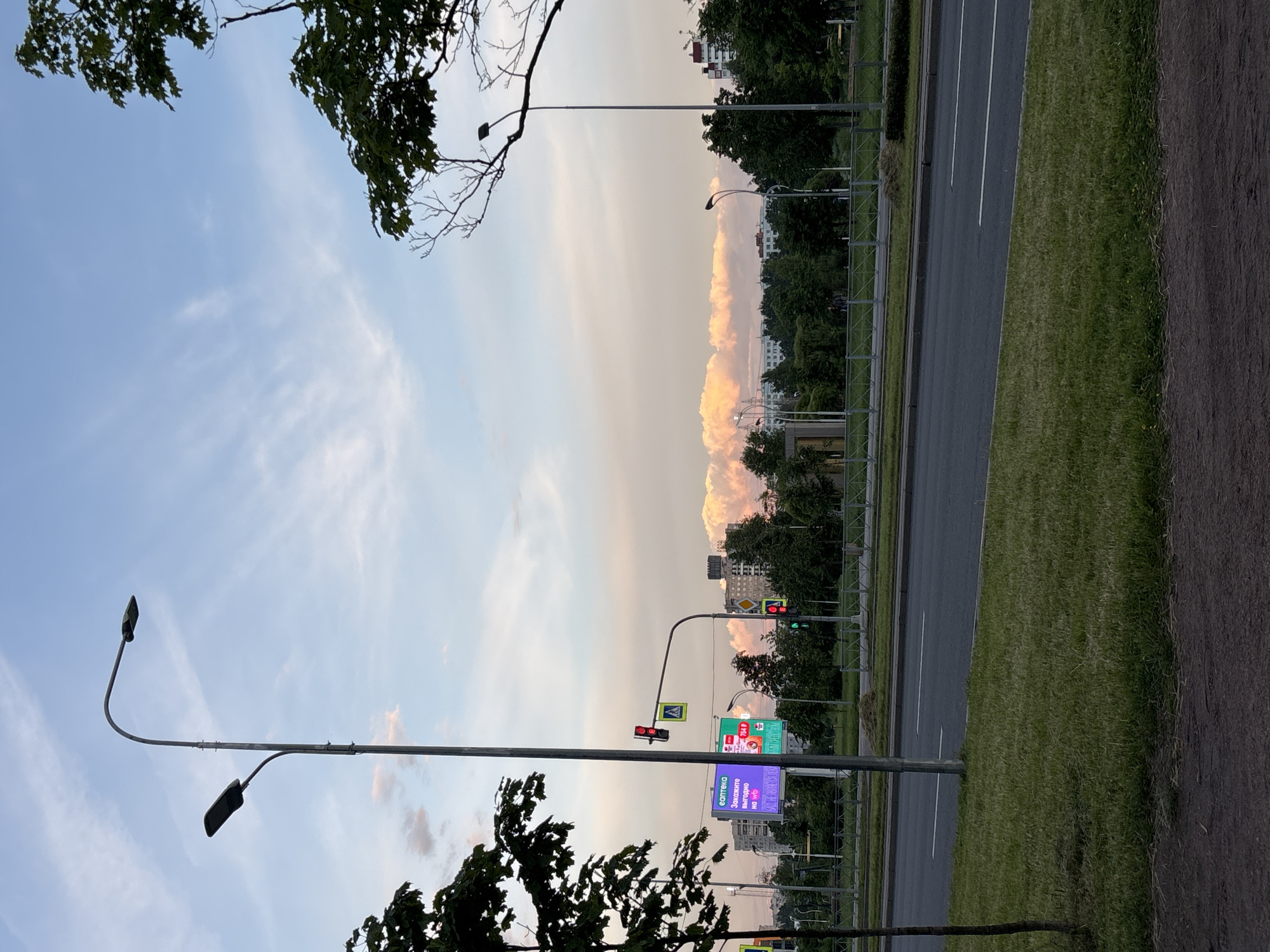 The image depicts an urban landscape with a road in the foreground and buildings and trees in the background. The sky is a pale blue with wispy clouds, some tinged with pink, suggesting either dawn or dusk. A tall street lamp with two lights stands prominently on the left side of the image, with another street lamp visible further down the road. Traffic lights and pedestrian crossing signs are present, indicating a road intersection. A large digital billboard is situated on the left, displaying colorful advertisements. Dense green trees line the area beyond the road, and several multi-story buildings are visible in the distance. The ground in the immediate foreground is a mix of asphalt and grass. There are no people clearly visible in the image.