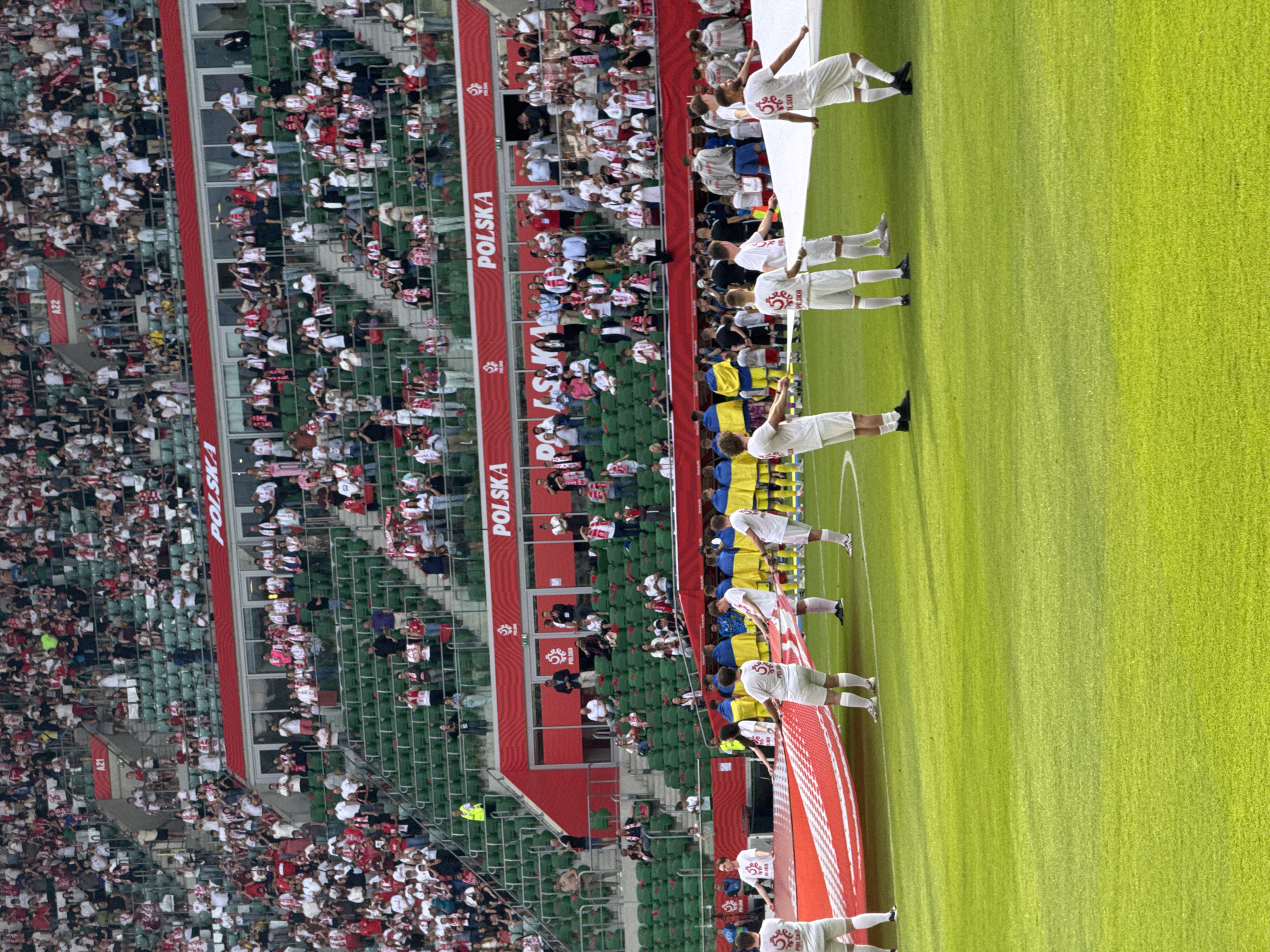 A soccer match unfolds in a stadium, with a large crowd filling the stands. The spectators are a mix of colors and shapes, a collective of fans from varying backgrounds. The scene is set against a backdrop of stadium architecture, marked by red signage proclaiming "POLSKA". On the field, teams in white uniforms are visible, their positions suggesting they are preparing for the match or in the midst of a ceremony. Patches with a white eagle design adorn the jerseys of some players. Some players wear blue and yellow jackets. The field is a vivid green. A partially visible red and white sign stretches across the foreground.