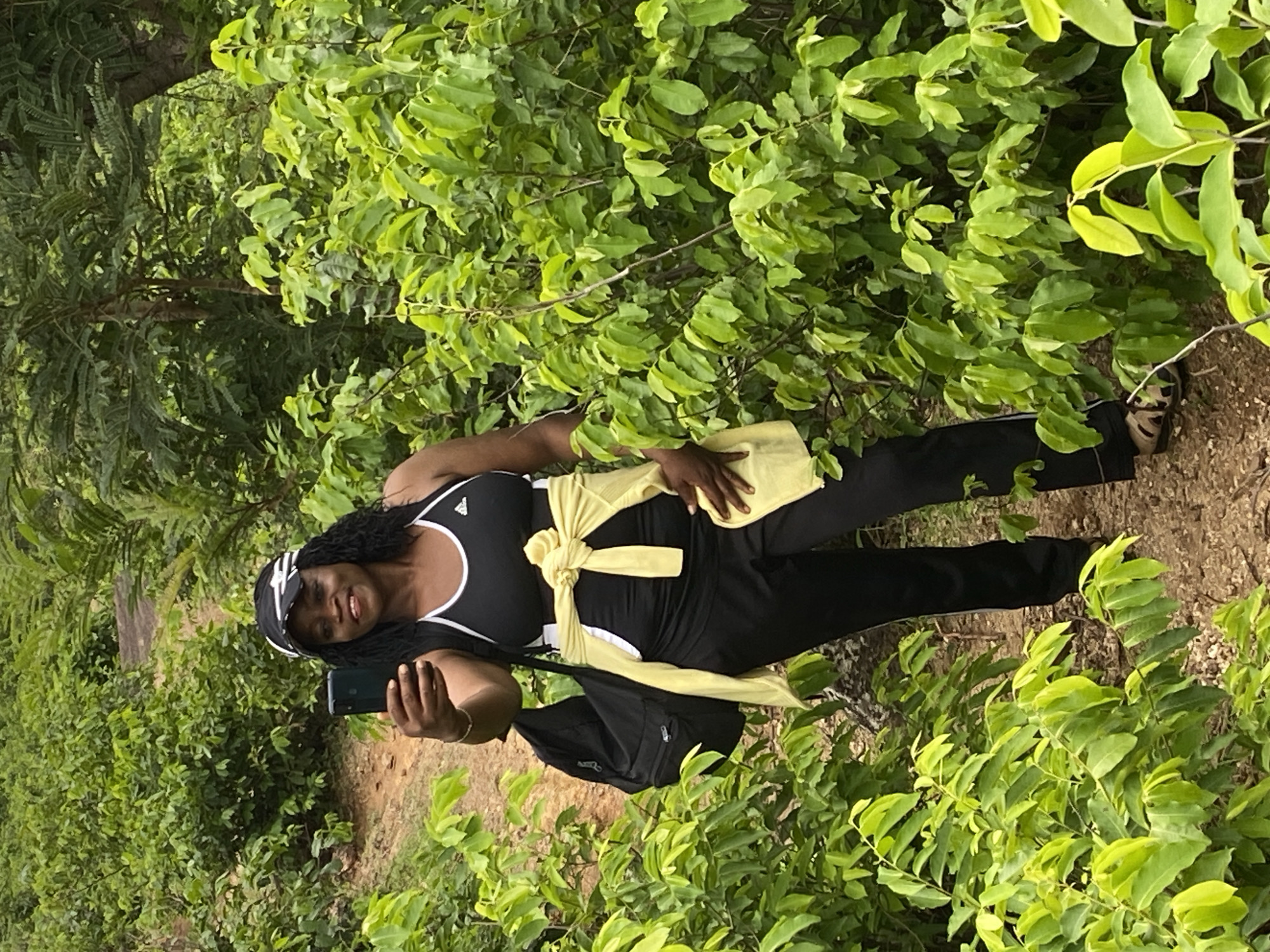 A middle-aged Black woman stands on a dirt path surrounded by lush green foliage. She is wearing a black and white tank top, black pants, and sandals. A yellow garment is tied around her waist. She is holding a smartphone in her left hand, presumably taking a selfie. A black backpack is slung over her shoulder. The location is Ganawuri, Nigeria.