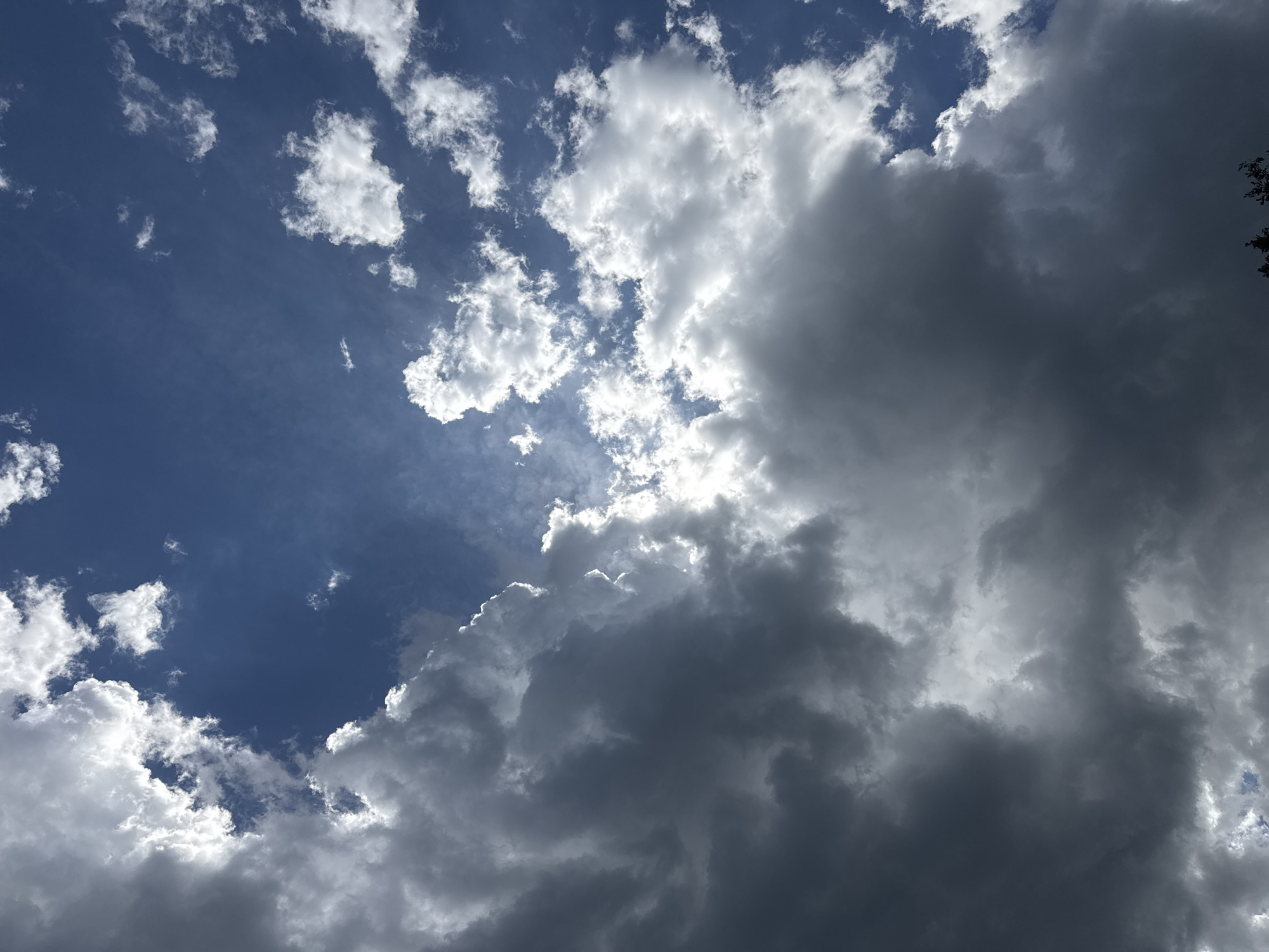 The image depicts a sky scene. A patch of deep blue sky is visible in the upper left quadrant, dotted with small, fluffy white clouds. To the right, a large mass of dark, ominous storm clouds dominates the view. The sun appears to be shining behind the clouds, creating a bright, diffused glow along their edges. The bottom part of the image consists of dark grey clouds. The image does not contain any recognizable location, people, subjects, objects, actions, interactions, or other environment of the city of Međuriječje, Montenegro.