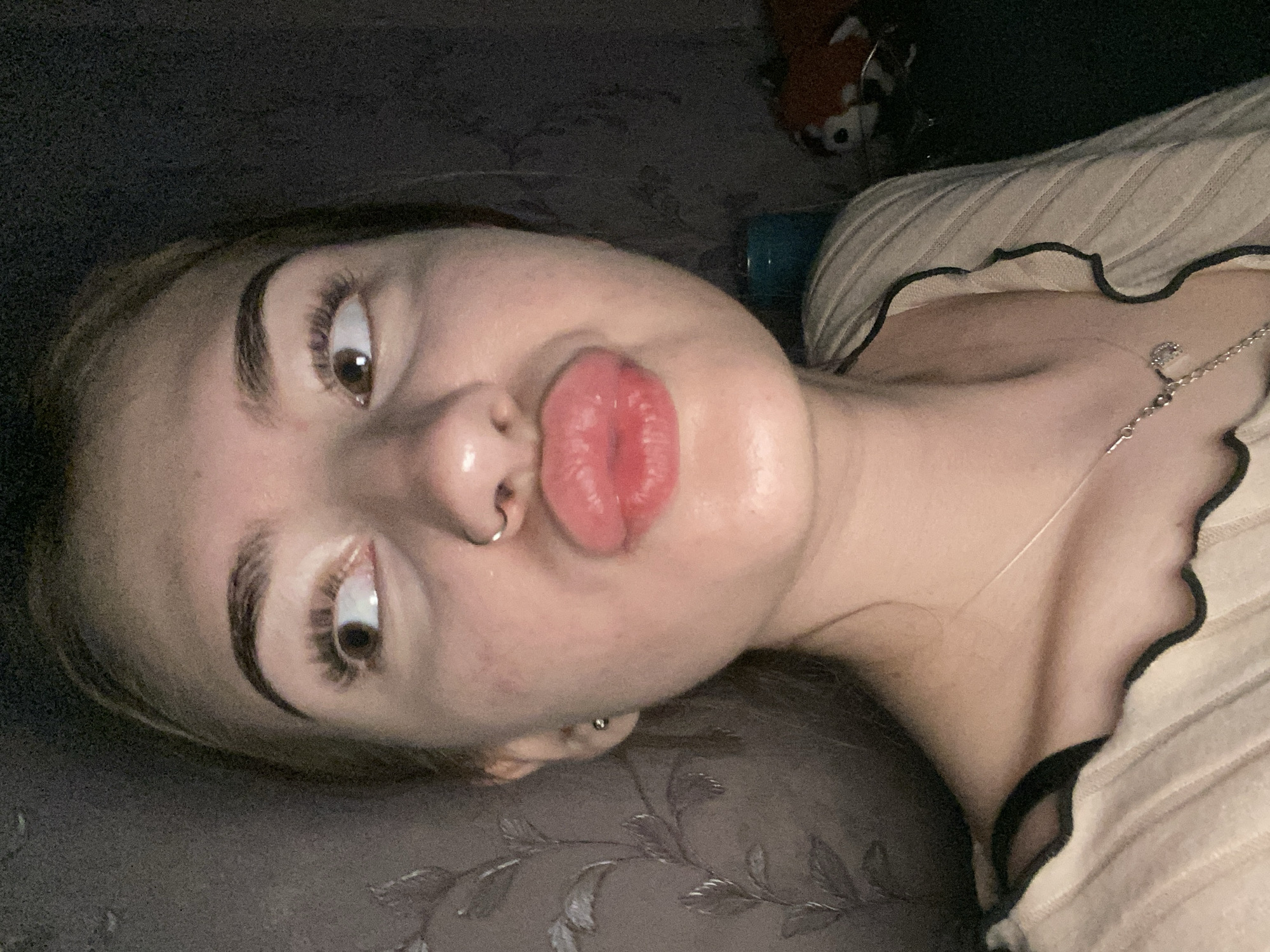A young woman with light brown hair is taking a selfie indoors. She is wearing a light-colored, ribbed top with black trim. She has a nose ring and is making a puckered face with her lips. Her eyes are looking to her right. The background appears to be a wall with a floral pattern and a dark, possibly wooden, surface. A stuffed animal is visible on the right side of the frame. The lighting is dim, suggesting it is nighttime or the room is dark. The location of Дьяково, Russia is not determinable from the image.