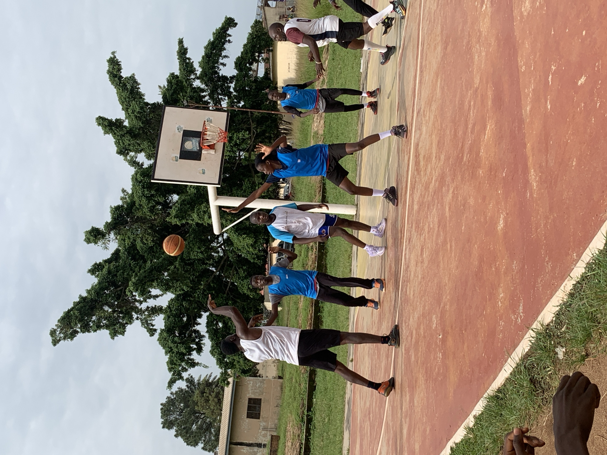 The image depicts a group of young men playing basketball on an outdoor court, possibly in Gura Top, Nigeria. The court surface is a faded red with white lines. In the background, there are trees with dense green foliage and a low-rise building with a brown roof. A basketball hoop with a white backboard and orange rim is visible. Six individuals are engaged in the game: one is shooting a basketball which is in mid-air, another is defending, and several others are positioned on the court. The players are wearing athletic attire, including shorts and jerseys, with some wearing bibs over their shirts. The weather appears overcast.