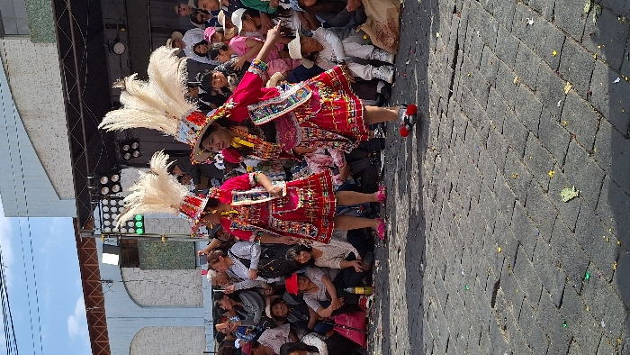 Two women, adorned in vibrant red dresses and tall white feathered headdresses, dance joyfully amidst a crowd. Their attire is rich with intricate patterns. The audience is mixed in age, some standing while others are seated. A stage-like structure is visible behind them, featuring stage lighting. The ground appears to be paved with dark gray brick. Sunlight streams down upon the scene, creating vivid contrasts.