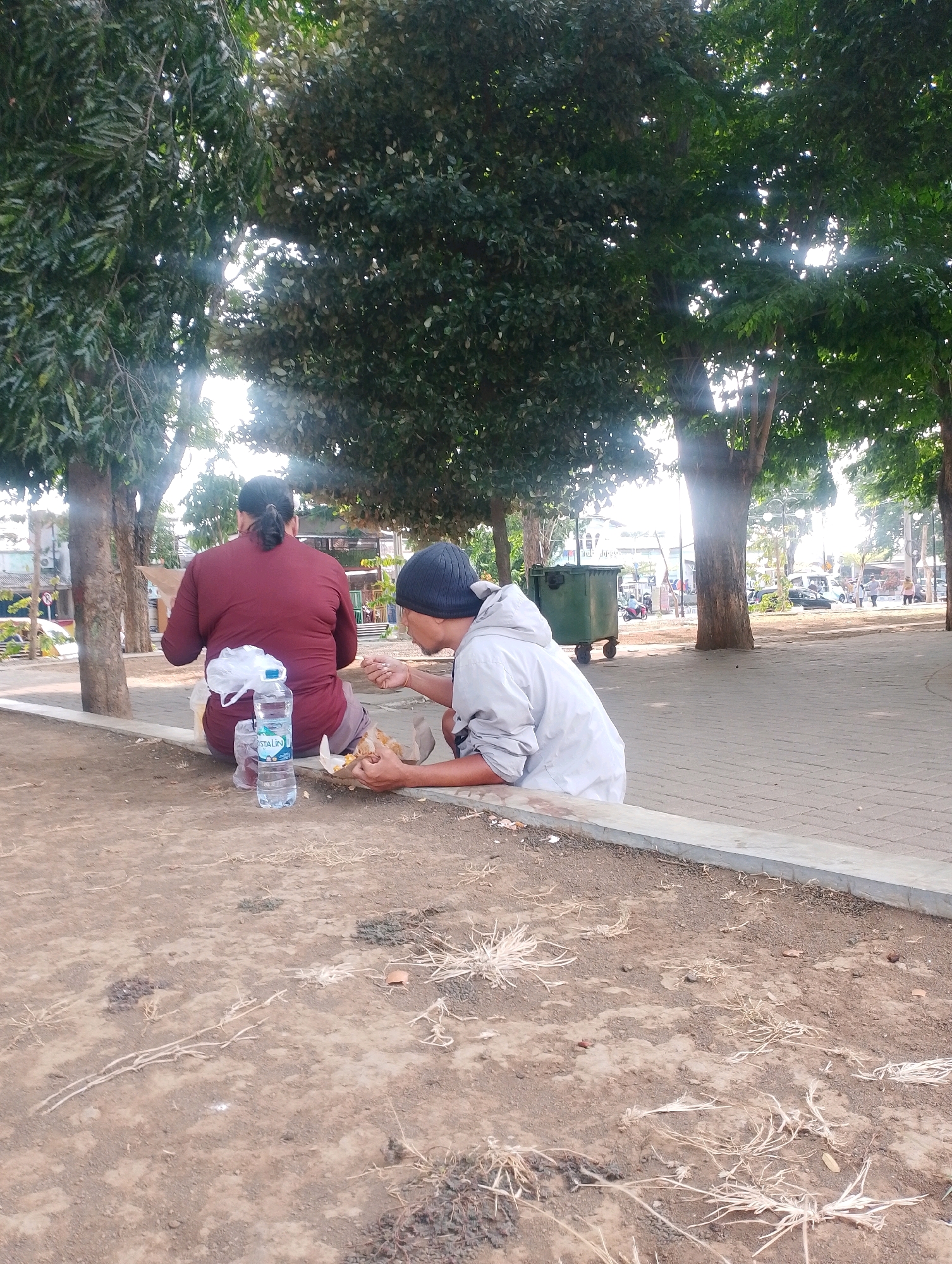 Two individuals are sitting on the ground in an outdoor urban setting, likely a park or public area, in Probolinggo, Indonesia. The person on the left, viewed from behind, wears a maroon long-sleeved shirt and has their hair pulled back in a bun. They are sitting next to a plastic bag and a bottle of water. The person on the right, facing away from the camera and towards the person on the left, wears a gray hooded jacket and a dark beanie. Both individuals appear to be eating food from paper wrappings.
The environment consists of a paved walkway or area with a raised concrete border in the foreground, leading to a dirt and gravel ground surface. Mature trees with dense foliage are present on the left and right sides of the frame, casting shadows and allowing sunlight to filter through their leaves, creating sunbeams. In the background, a green trash bin on wheels is visible, along with a street with light traffic, including cars and motorcycles. Buildings and streetlights can also be seen in the distance, indicating a city environment. The overall scene suggests a casual moment of rest and eating in a public space.