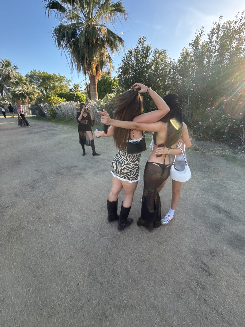 A group of young women is standing on a dusty ground, likely in the city of Indio, United States, given the presence of palm trees and other desert vegetation. The woman closest to the camera is wearing a zebra-print skirt, a black top, and black boots. Another woman is wearing a sheer black dress. They have their arms around each other. Another woman is standing a short distance away, pointing something towards them while holding a camera. Other people and palm trees are visible in the background.