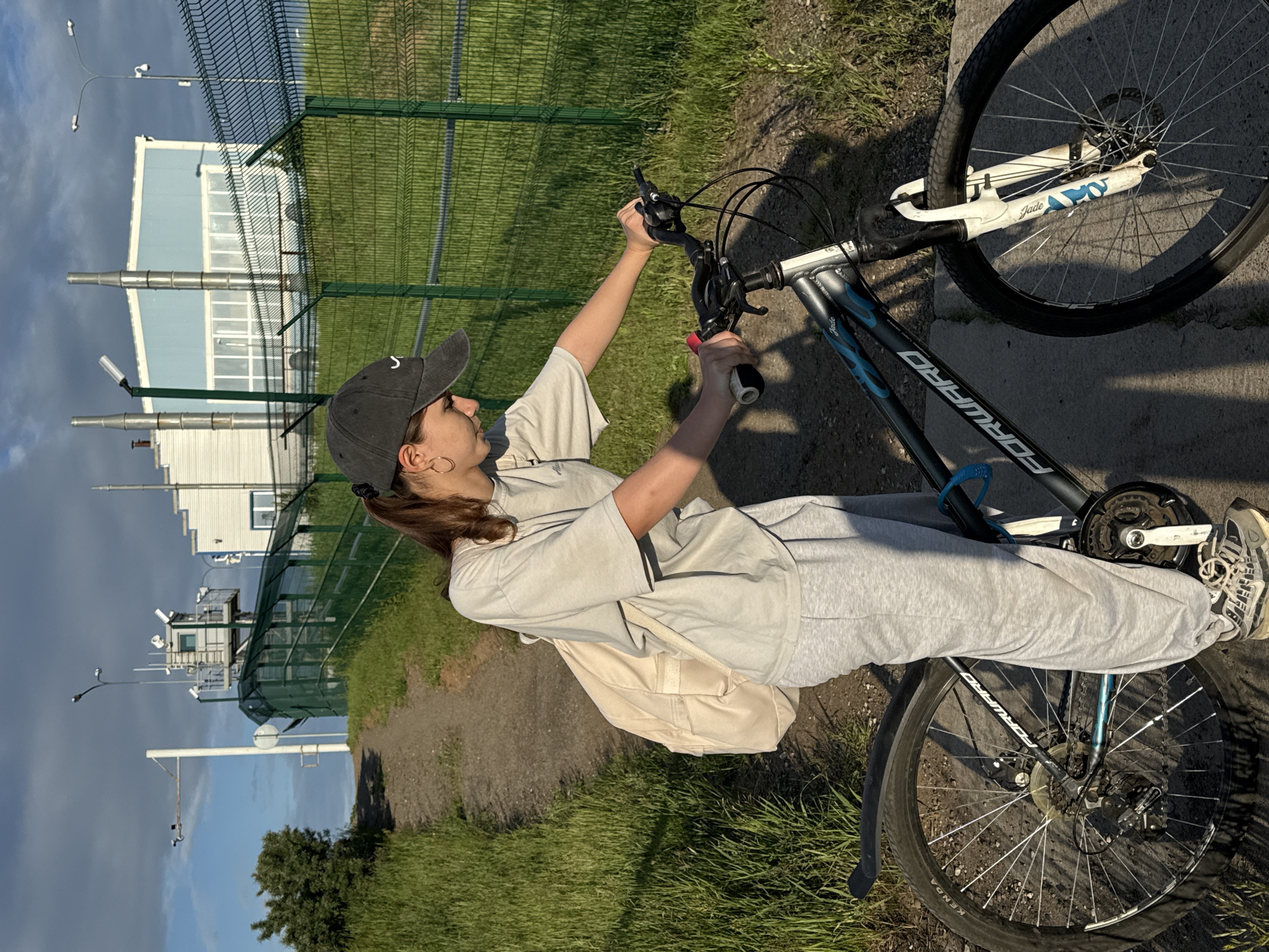 The image depicts a young woman standing beside a bicycle on a dirt path in Chaikovsky, Russia, at what appears to be an industrial or secured area. The woman, positioned to the right of the bicycle, wears a dark baseball cap, a light grey oversized t-shirt, light grey sweatpants, and sneakers. She carries a light beige backpack on her left shoulder and has a ponytail visible from beneath her cap. Her hands are on the handlebars of a dark grey mountain bike, which features "FORWARD" branding and blue "Jade" accents on the frame and white front fork. The bicycle is equipped with disc brakes and Kenda tires.
The path, composed of dirt and gravel, ascends gently from the lower left towards the upper center-left. Green grass and scattered weeds line both sides of the path. To the right of the path, long shadows are cast on the ground, indicating direct sunlight from the left.
A tall, dark green mesh fence runs parallel to the path and grassy embankment in the midground, separating the path from the structures beyond. Behind this fence, on an elevated position, a multi-story security observation post or control tower is visible, featuring a light grey structure with white upper sections, multiple windows, and various cameras and communication devices affixed to its exterior. Further to the right and in the background, several light blue and white industrial-style buildings with numerous windows and prominent vertical metallic pipes or chimneys are present. Two streetlights are also visible within this area. To the far upper left, a white pole with two mounted security cameras is visible against a partly cloudy sky, which shows patches of blue.