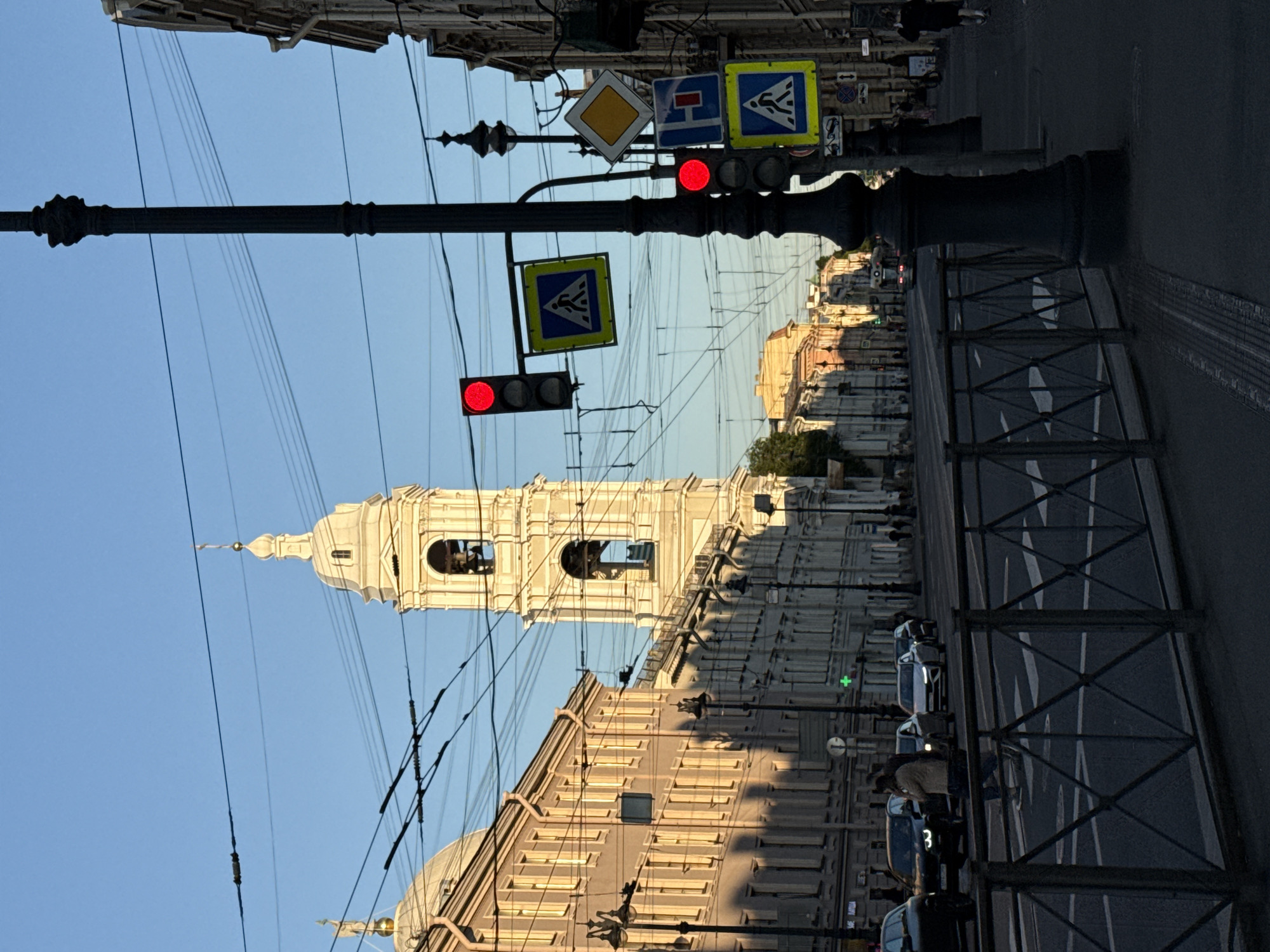 This image depicts a street scene in Saint Petersburg, Russia, with the prominent bell tower of the Vladimirskaya Church (Vladimir Cathedral) visible in the background. The time of day appears to be late afternoon or early evening, characterized by a clear blue sky and long shadows cast from the buildings on the left side of the street across the road.
In the immediate foreground, a dark metal fence with a diamond lattice pattern runs horizontally, separating the sidewalk from the vehicular lanes. On the left side of the road, two cars are visible: a dark SUV with illuminated headlights and a lighter-colored sedan. Two pedestrians are centrally positioned, walking across a white-striped crosswalk from left to right. The individual on the left wears a light-colored jacket, while the one on the right is partially obscured.
On the right side of the foreground, a dark, ornate streetlamp pole stands vertically, partially obscuring various street signs and a traffic light. Attached to this pole are two red circular traffic lights, both illuminated, and several road signs. These signs include two blue square signs with white pedestrian symbols, a yellow diamond-shaped sign with a white border, and a blue rectangular sign with a red horizontal bar. Further down the right sidewalk, another similar streetlamp is visible, along with multiple pedestrians.
The street is a multi-lane thoroughfare with visible white lane markings. Multiple vehicles, including cars and a trolleybus or tram in the distant background, are present on the road.
Buildings line both sides of the street. On the left, a multi-story edifice with a light-colored facade features numerous rectangular windows and a domed section visible on its far left. This building is illuminated by direct sunlight on its upper sections. Below it, a green cross symbol, likely indicating a pharmacy, is visible on a shopfront. Trolleybus overhead lines are strung across the street. The buildings on the right side of the street are largely in shadow, exhibiting classical architectural details. The bell tower of the Vladimirskaya Church, a multi-tiered white structure with archways for bells, stands prominently at the end of the street in the distance, illuminated on its right side by the setting sun.