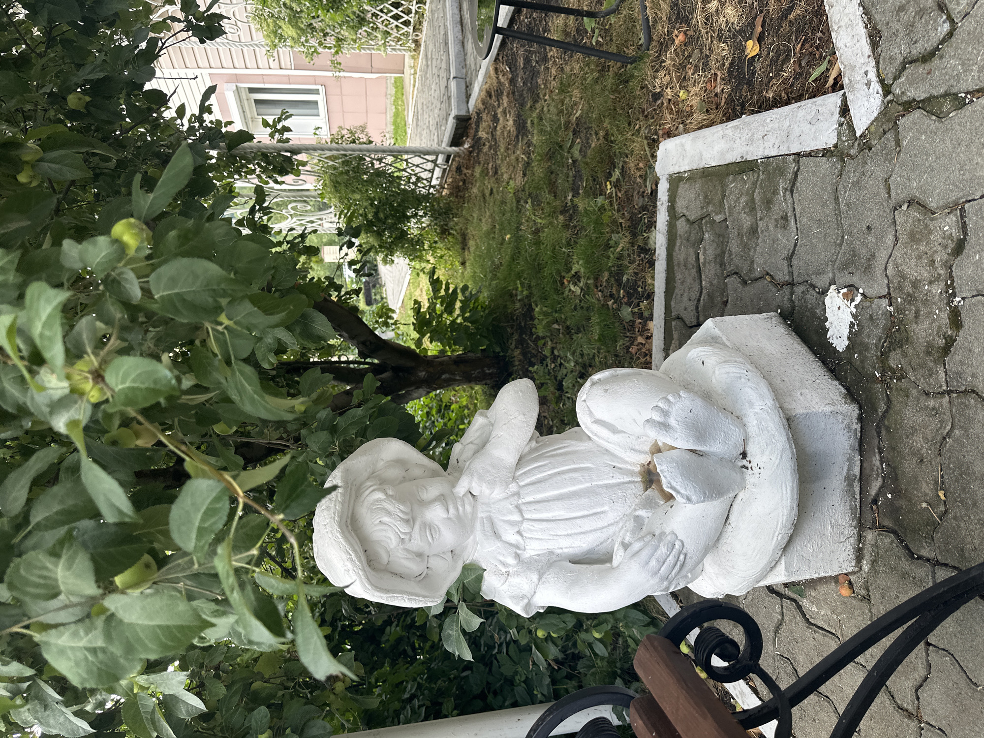 A white plaster garden statue of a young child wearing a wide-brimmed hat and a ruffled dress is seated on a raised concrete pedestal in what appears to be a backyard or garden area. The child is depicted with one finger touching its chin, in a thoughtful pose. The statue is positioned on interlocking concrete pavers, with a dark-colored park bench visible to the left. Behind the statue, lush green foliage from a tree with small green fruits hangs overhead, with a building with light pink siding and a white window visible in the background. A white decorative metal fence separates the paved area from a grassy patch. The environment suggests a residential outdoor space in the city of Danilovka (Altai), Russia.