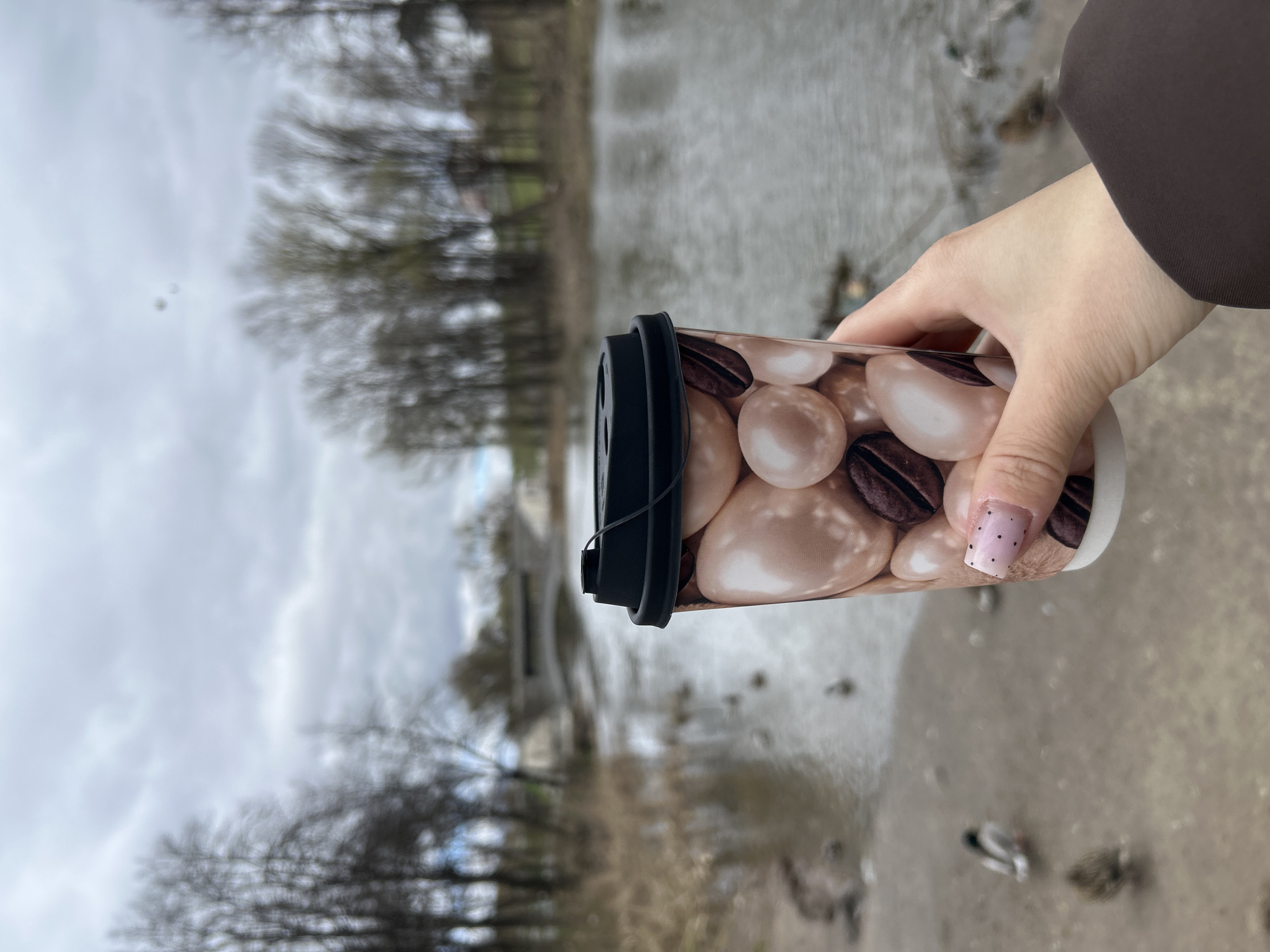 A human hand holds a disposable coffee cup in an outdoor setting in Klin, Russia. The hand, wearing a brown long-sleeved garment, has light pink nail polish with small black polka dots on the visible fingernail. The coffee cup features a decorative pattern of brown coffee beans interspersed with smooth, light beige, oval-shaped candies or stones, and has a black plastic lid.
The background shows a wide body of water, likely a river or lake, stretching into the distance. Several dark-feathered ducks or small birds can be seen on the water's surface towards the lower right. The far banks of the water body are lined with numerous bare trees, silhouetted against an overcast, cloudy sky, suggesting a cold season like late autumn or winter. The overall lighting is soft and diffused, indicative of a daytime scene under heavy cloud cover. The image captures a serene moment of someone enjoying a beverage while outdoors by the water. There is no visible text.