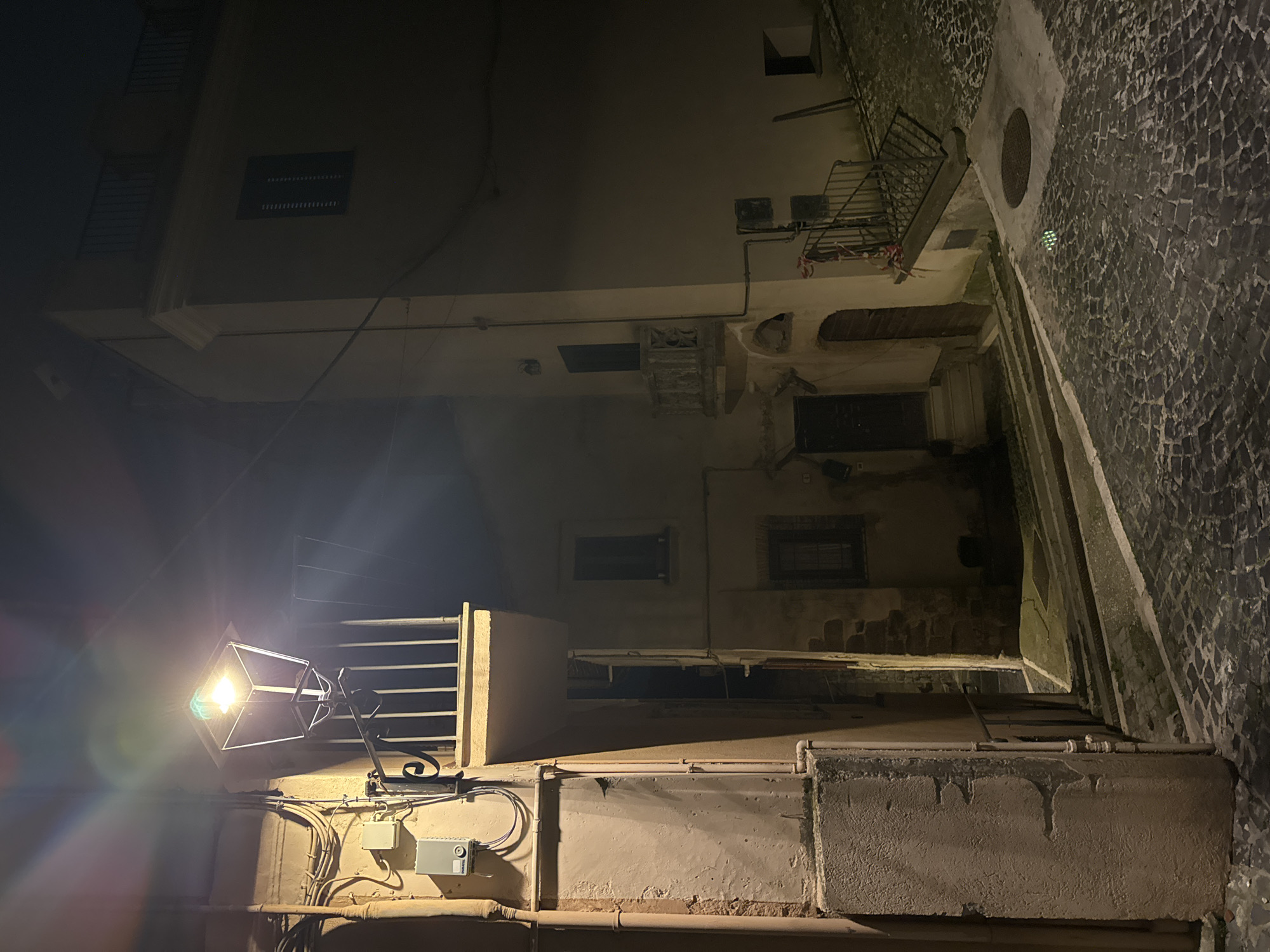 The image shows a narrow street in Castelnuovo di Porto, Italy, at night. The street is paved with dark cobblestones. Buildings line the street, constructed of light-colored stone or stucco. Windows are visible on multiple levels. A streetlight illuminates the lower portion of the street and building walls, casting a bright glare. Plumbing pipes and electrical conduits are visible on the surface of the lower walls. A manhole cover is embedded in the cobblestone street surface. There are stairs in a passageway. Balconies are attached to the buildings.