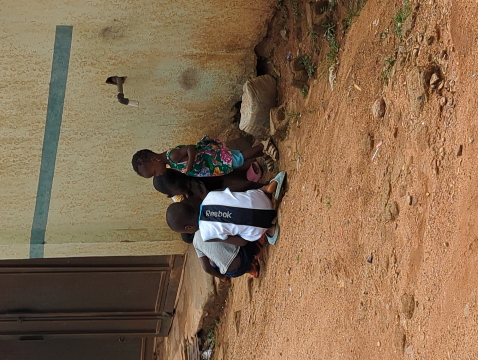 The image shows a group of children near a building in Sabon Tasha, Nigeria. The building has a light-colored wall, a door, and a pipe extending from the wall. The ground is dirt and gravel. The children are clustered together, engaging in an activity. One child is wearing a Reebok shirt, another a floral dress.