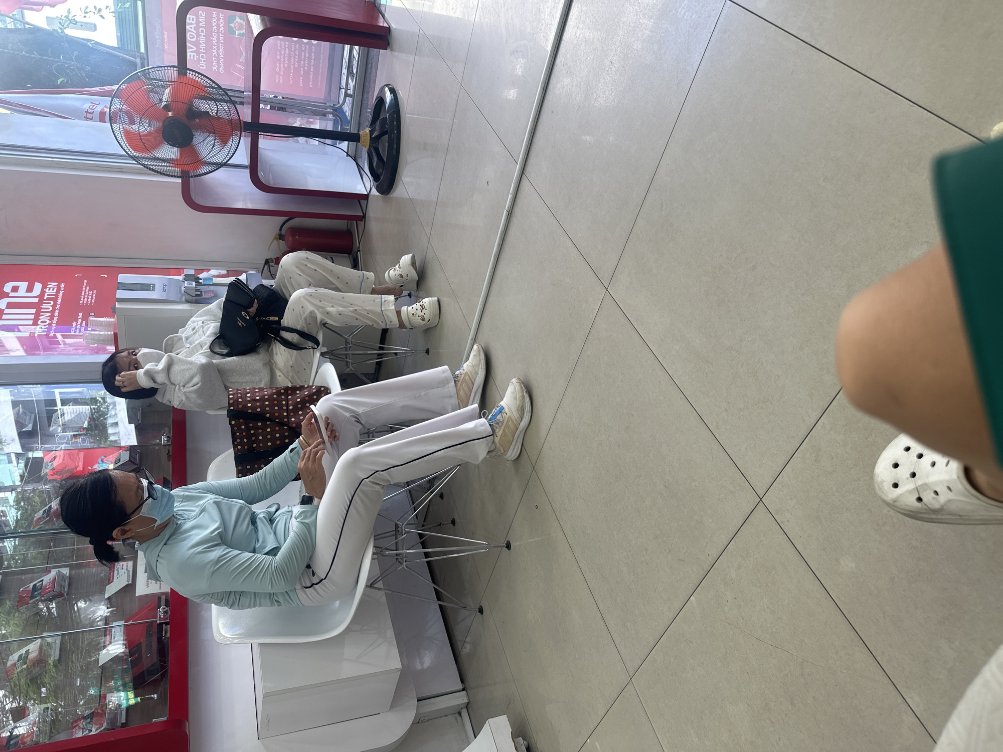 The image captures an indoor waiting area, likely a customer service center or shop in Nha Trang Ward, Vietnam, during daytime. Two women are seated, while the photographer's leg and foot are visible in the foreground.
On the left, a woman wearing a light blue long-sleeved top, white pants with black side stripes, beige sneakers, and a light blue face mask is seated, looking down at a mobile device held in her hands. To her right, another woman, also wearing a face mask, a light-colored top, and patterned pants, is seated. She has a black handbag resting on her lap and is looking down. She wears white platform Crocs-style shoes. The person taking the photo is wearing a green garment and white Crocs-style shoes.
The setting includes a light-colored tiled floor with visible grout lines. A red metal frame structure runs along the wall, possibly for displays or as a room divider. A large black pedestal fan with orange blades is prominently placed near the window, turned on and providing air circulation. Behind the fan, a red fire extinguisher is visible. A water dispenser with cups is also seen in the background. Large windows reflect parts of the outdoor environment and the interior space.
Visible text on red signs includes "TRỌN ƯU TIÊN" (Complete Priority/Full Privilege) and "Dịch vụ di động dành cho khách hàng ưu tiên" (Mobile service for priority customers), indicating a customer-oriented business. Other partial and inverted text, such as "ttel", "doup", "Jettelv", "mooslat", "VOAS", and "DUHT DAX VÃO DUDUH", is present on the window reflections and signs, further confirming the Vietnamese setting. The overall atmosphere appears calm and routine, with individuals waiting patiently.