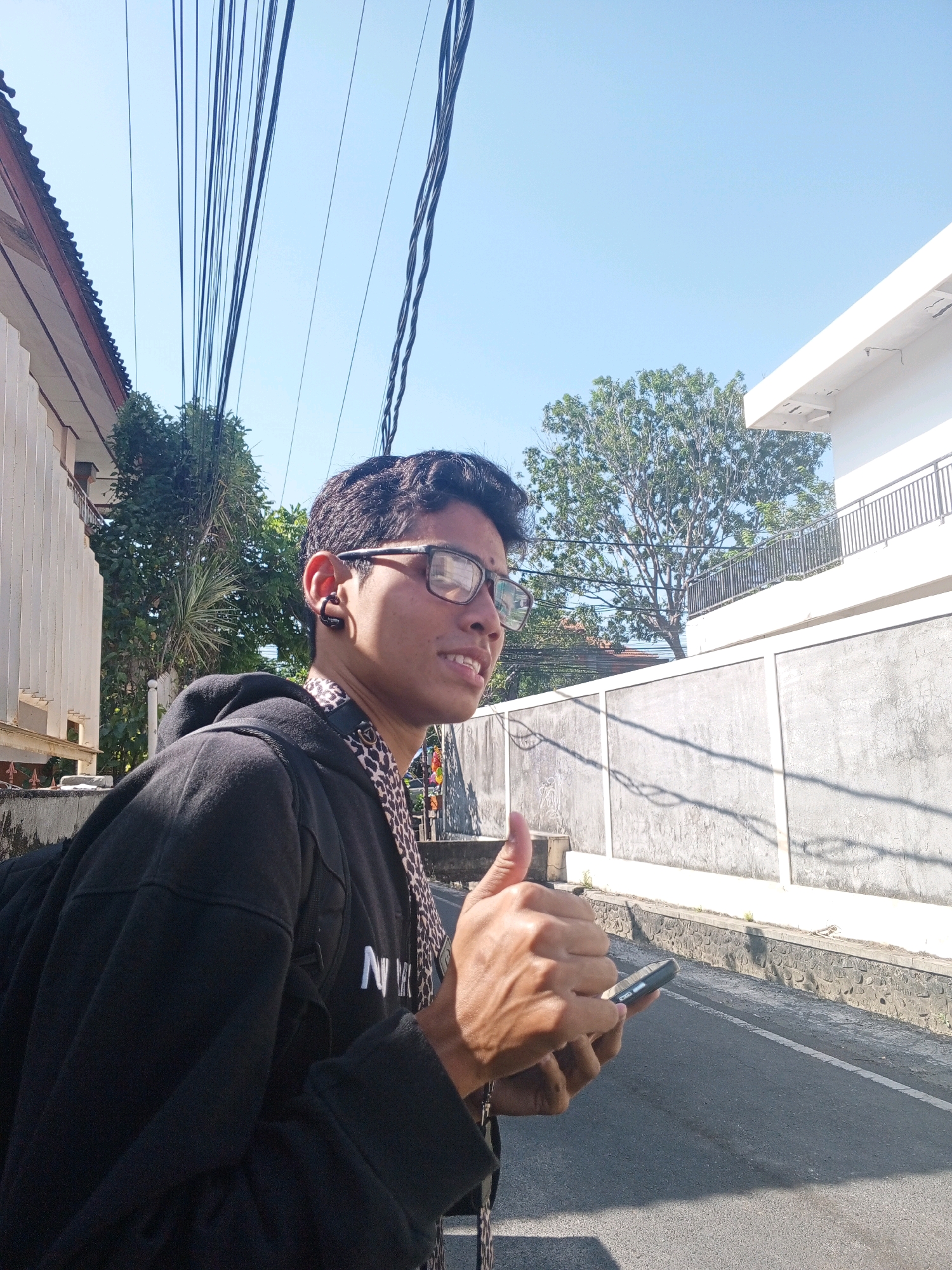 An over-the-shoulder medium shot captures a young man in Denpasar, Indonesia, standing outdoors and giving a thumbs-up while holding a smartphone. He is looking to his right, away from the camera, with a slight smile. The man wears glasses and a black hooded jacket over a leopard-print shirt. He has a backpack slung over his left shoulder and an ear piercing on his right ear. The background reveals a street with a textured gray concrete wall and a building with a white facade and railing. Overhead, a tangle of black electrical wires crisscrosses the clear blue sky. Sunlight casts shadows on the street and wall, indicating a bright, sunny day. The environment suggests an urban setting, consistent with Denpasar.