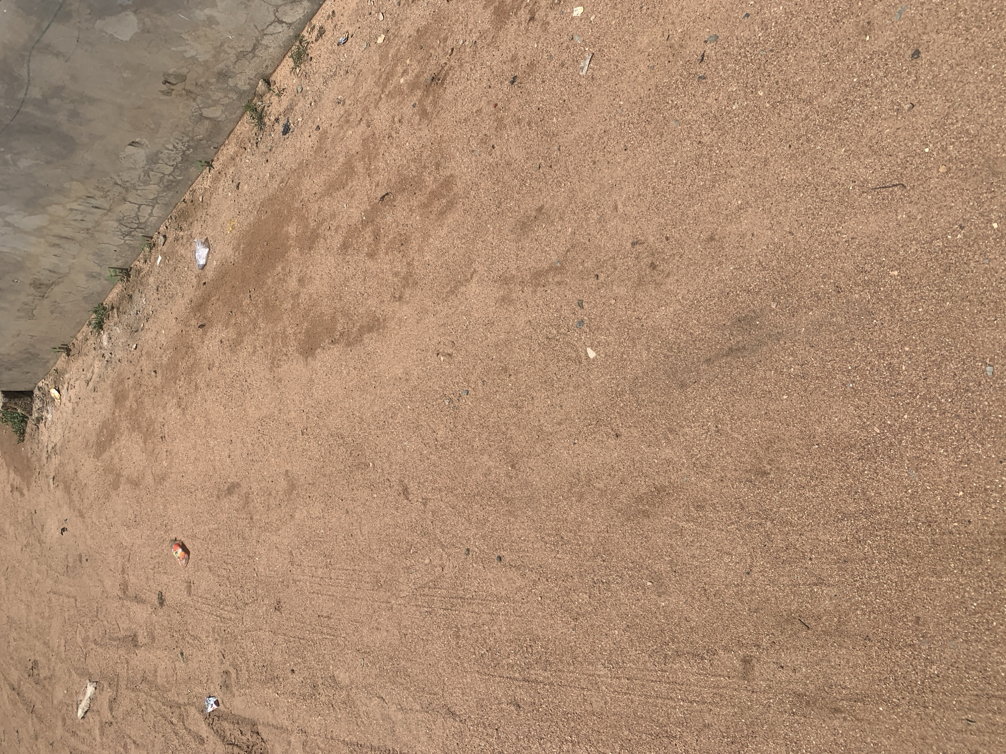 The image depicts an outdoor scene with a sandy ground surface. A concrete wall runs along the right side of the frame. Scattered on the ground are various small objects, including a piece of orange and red colored plastic, a crumpled white wrapper, and a few small stones. There are also some sparse green plants growing along the base of the wall. The sandy ground shows some tire tracks and appears to be somewhat uneven. The overall impression is that of an unpaved area possibly in a less developed part of a city. This scene is located in Jauro Yinu, Nigeria.