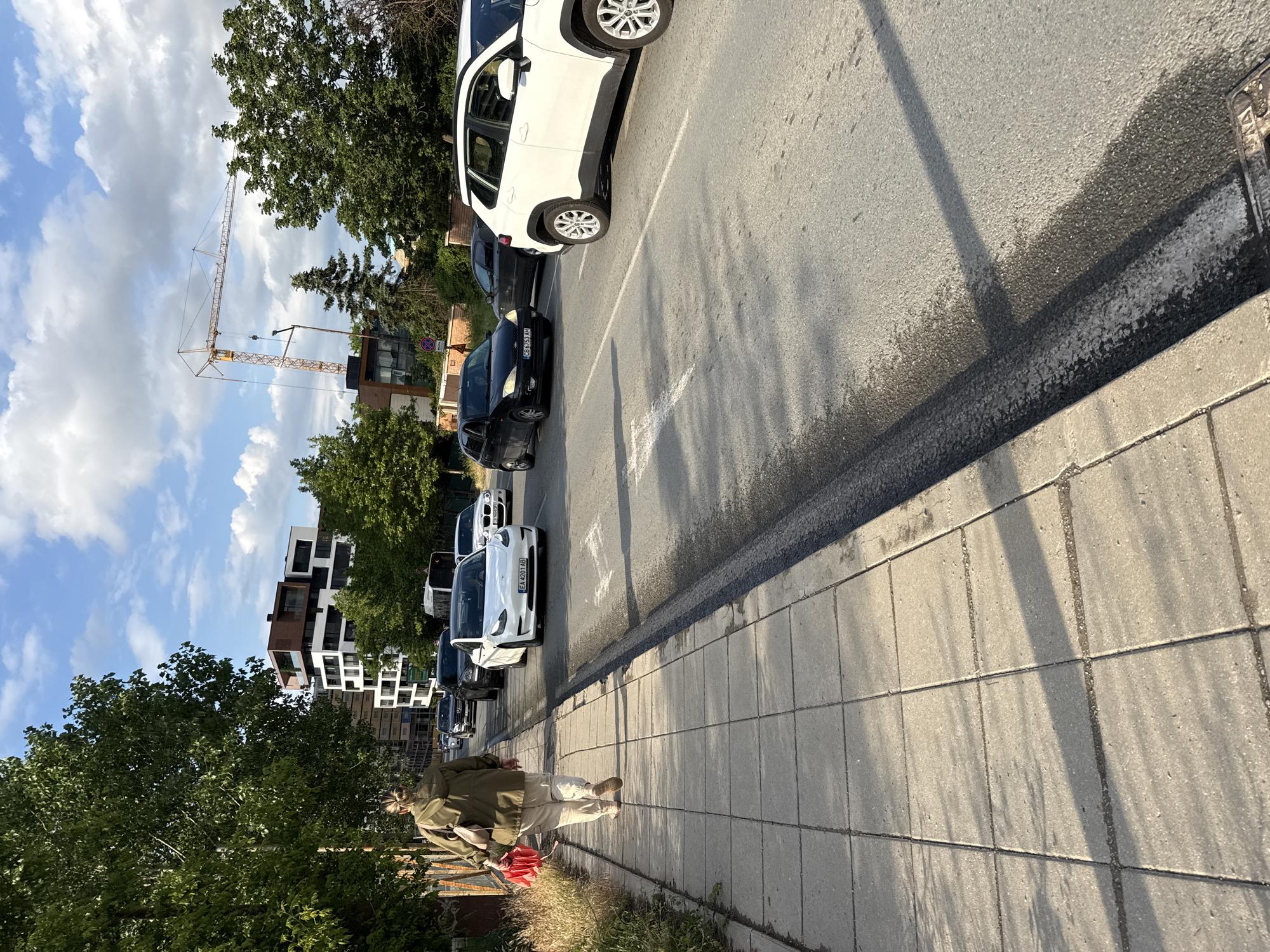 This image depicts an urban street scene, likely in Sofia, Bulgaria, captured during what appears to be the late afternoon or early evening due to the long shadows cast by the sun. The sky above is bright blue with scattered white cumulus clouds.
On the right side of the frame, a paved sidewalk with square concrete slabs extends into the distance. A person, viewed from behind, walks along this sidewalk. They are dressed in a light green or khaki jacket, light-colored trousers, and shoes, and are carrying a red, folded object, possibly an umbrella or bag.
To the left of the sidewalk is an asphalt road with faded white parking lines. A line of cars is parked along the curb. Prominently featured are a white SUV, followed by a black sedan displaying the Bulgarian license plate "CB 5313 H". Further down the street, another white sedan, possibly an electric car, is visible with the Bulgarian license plate "EA 4201 AB". A traffic sign indicating "No Stopping" (a red circle with a blue diagonal line) is partially visible near the parked cars.
In the background, behind the vehicles and trees, modern multi-story buildings with distinct architectural features, including white, black, and brown paneling, stand. A large yellow construction crane dominates the left side of the background, suggesting ongoing development. The scene is rich with green foliage from various trees and shrubs lining the street. The sunlight creates strong contrasts and highlights the textures of the pavement and road.