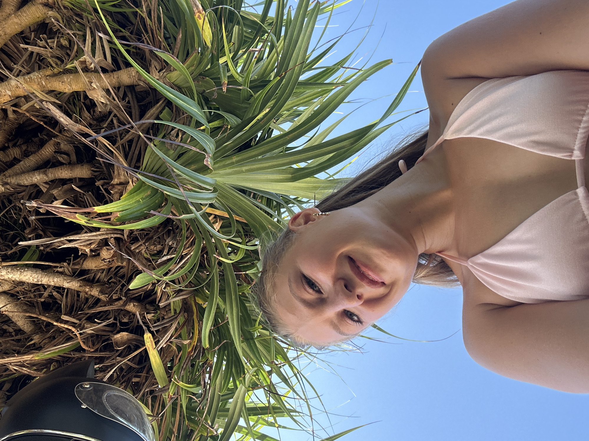 The photo is taken from a low angle, looking up at a young woman standing in front of a screwpine tree, likely in Kuta Selatan, Indonesia. The woman is fair-skinned with brown hair and is wearing a light pink garment. She is smiling and looking down at the camera. Parts of a black object, possibly a motorcycle helmet, are visible at the bottom left. The background consists of a clear blue sky. The image is taken outdoors and suggests a sunny day.