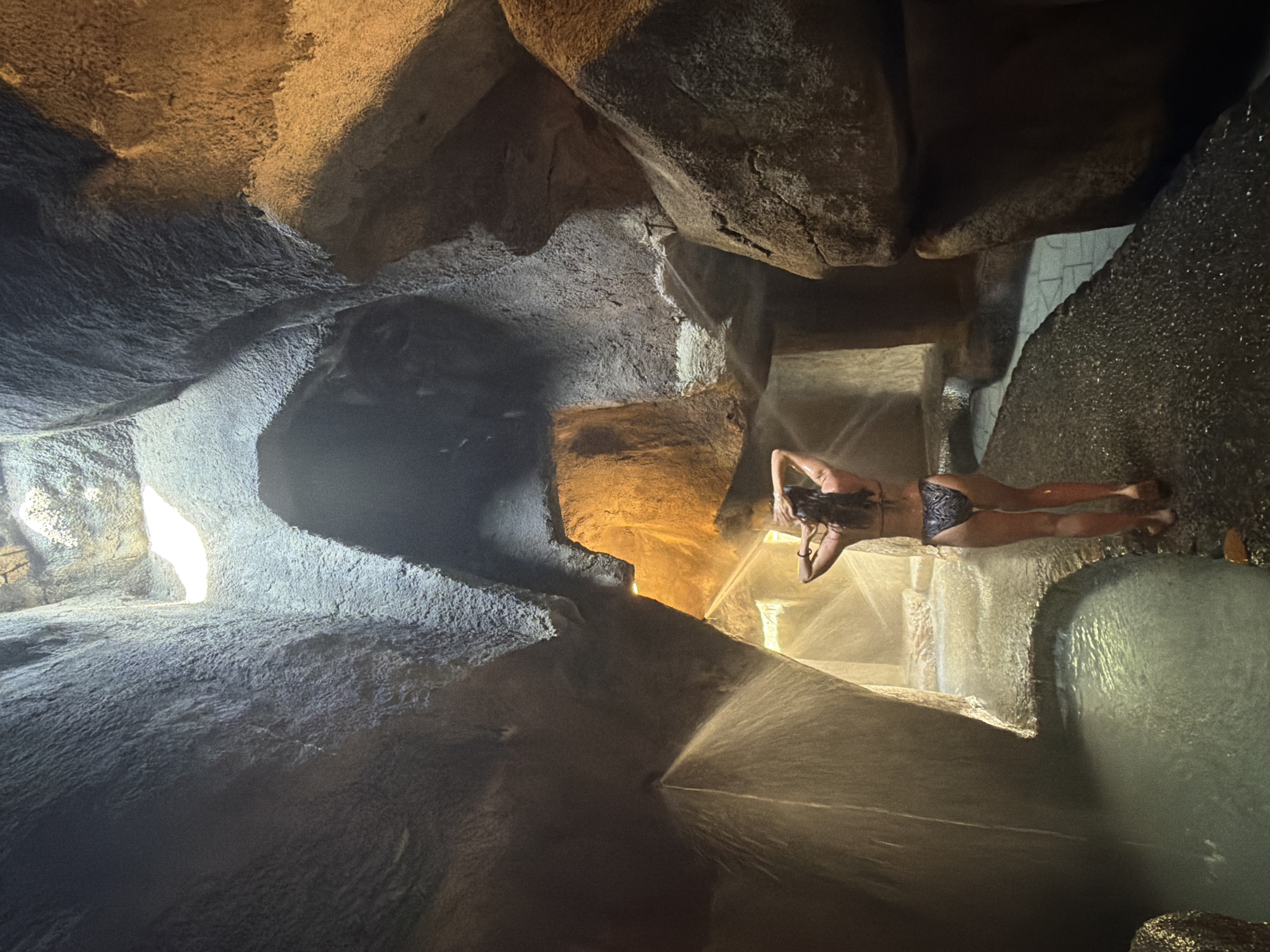 A woman in a black and white patterned bikini stands with her back to the camera in a naturalistic grotto setting. She has her hands on her head, with her wet hair cascading down her back. Water is spraying from above and from the sides of the grotto, creating a misty atmosphere. The grotto walls appear to be made of rough, uneven stone, with openings that allow light to filter in from above. A pool of water is visible in the foreground, and a wet, textured pathway leads around it. The overall impression is that of a natural hot spring or spa environment. While the specific location in Tay Nha Trang Ward, Vietnam is not visible in the image, the setting is suggestive of a tropical, geothermally active area.