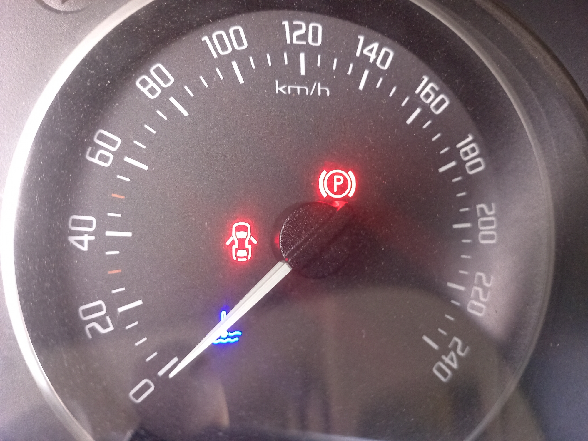 The image shows a close-up of a car's speedometer. The speedometer displays speed in kilometers per hour, with markings for 20, 40, 60, 80, 100, 120, 140, 160, 180, 200, 220, and 240. There are three illuminated warning lights on the dashboard: a red brake warning light with a "P" symbol inside a circle, a red warning light depicting an open car door, and a blue warning light with wavy lines, likely indicating low coolant or engine temperature. The needle of the speedometer is pointing between 20 and 40 km/h, closer to 20 km/h. The environment appears to be the interior of a vehicle, and there are no people visible. The image quality is slightly blurry and dusty, suggesting it was taken inside a car, possibly parked or moving at a low speed. There is no information within the image to determine its location in Novokuznetsk, Russia.