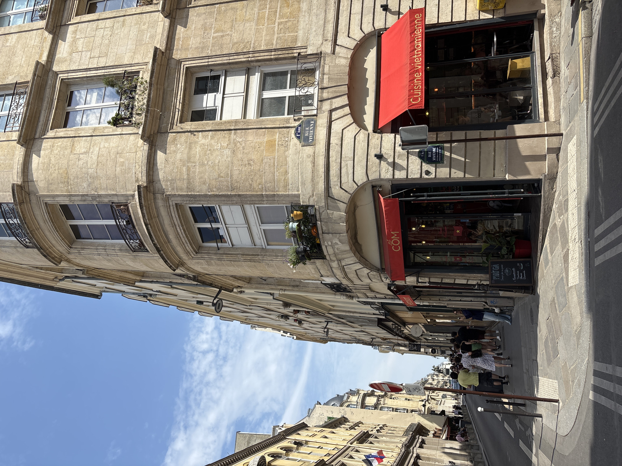 The image shows a street scene in Paris, France, likely in the 1st arrondissement given the street sign visible which reads "1er Arr. Rue Vivienne". The perspective is from a sidewalk looking down a street lined with buildings. On the right, a corner building features a restaurant with a red awning that says "Cuisine Vietnamienne" and another red awning above it with "COM". The building is made of stone with multiple windows, some with balconies. On the left side of the street, there are other buildings, flags, and a street with cars and pedestrians. Several people are walking down the sidewalk, some are facing away from the camera. The sky is blue with some clouds. The overall scene depicts a typical sunny day in a Parisian street.