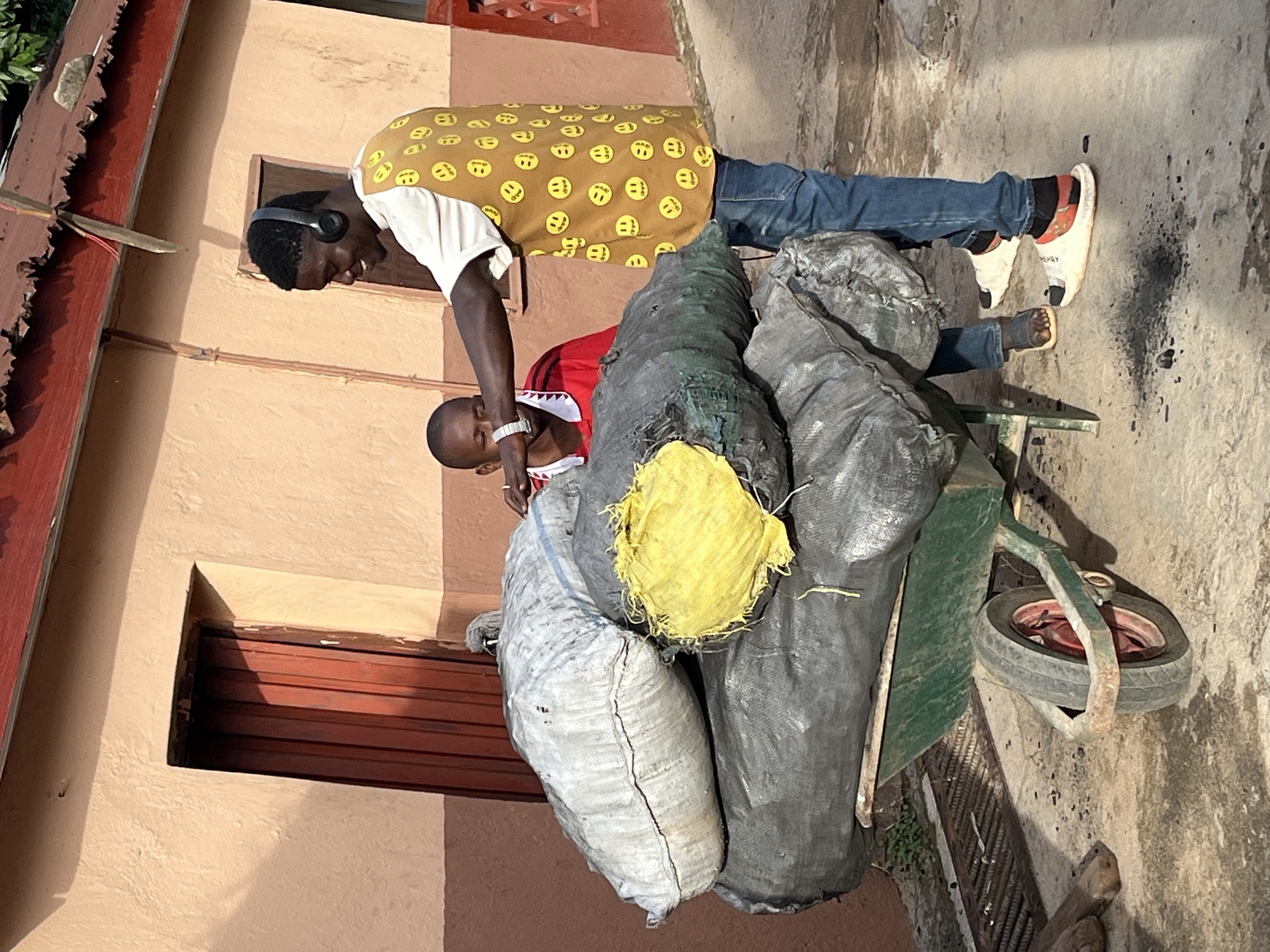 Two men are depicted outdoors, engaged with a loaded handcart next to a building in Jos, Nigeria. The scene is brightly lit by natural sunlight, casting distinct shadows.
The man on the left, standing, has dark skin and is wearing a white t-shirt, blue jeans, and a mustard yellow apron covered in small yellow smiley face emojis. He has black over-ear headphones and white sandals with dark socks. He is leaning forward over the handcart, looking down, seemingly inspecting or adjusting the contents.
The second man, closer to the right and bending over, also has dark skin. He wears a red short-sleeved shirt with a white collar that features a shark-tooth pattern, and dark trousers or shorts. A silver watch is visible on his left wrist. He appears to be interacting with the handcart or its contents.
The handcart, featuring a green frame and a single wheel with a red rim, is heavily loaded with large, bulky bags. These include several grey or black sacks, one white sack, and a small piece of yellow fabric covering part of one black bag. The contents of the bags appear to be a dark, granular material, possibly charcoal or another bulk commodity.
The background shows a light brown or tan building with a dark red/brown window shutter and a matching roofline. The ground is a rough concrete or dirt surface with some dark splatters. The overall activity suggests the men are involved in transporting or handling goods using the handcart.