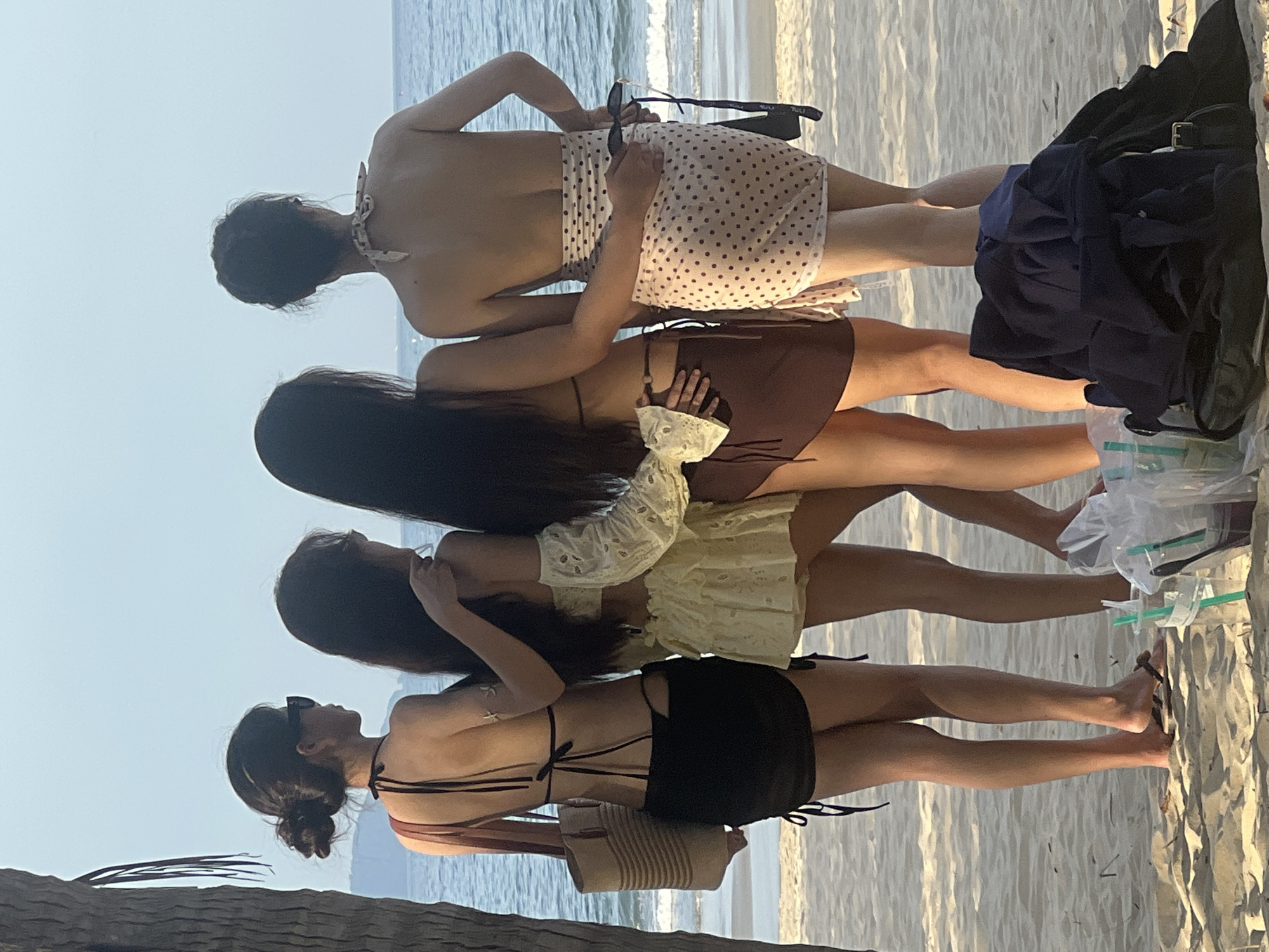 Four individuals stand on a sandy beach facing the ocean. The two individuals on the left are wearing black and yellow swimwear, respectively, and have their arms around each other. The person on the far left carries a straw bag. The two individuals on the right are wearing bikinis and sarongs. One person on the right is wearing a brown sarong and the other a polka-dot sarong. All individuals appear to be adults. The environment is a beach with a tree on the left, sand in the foreground, and the ocean and sky in the background. There are plastic cups and bags on the sand in the foreground. The image is taken from behind the individuals, looking towards the ocean. This scene is consistent with a beach location in Đà Nẵng, Vietnam, a popular coastal city known for its beaches.