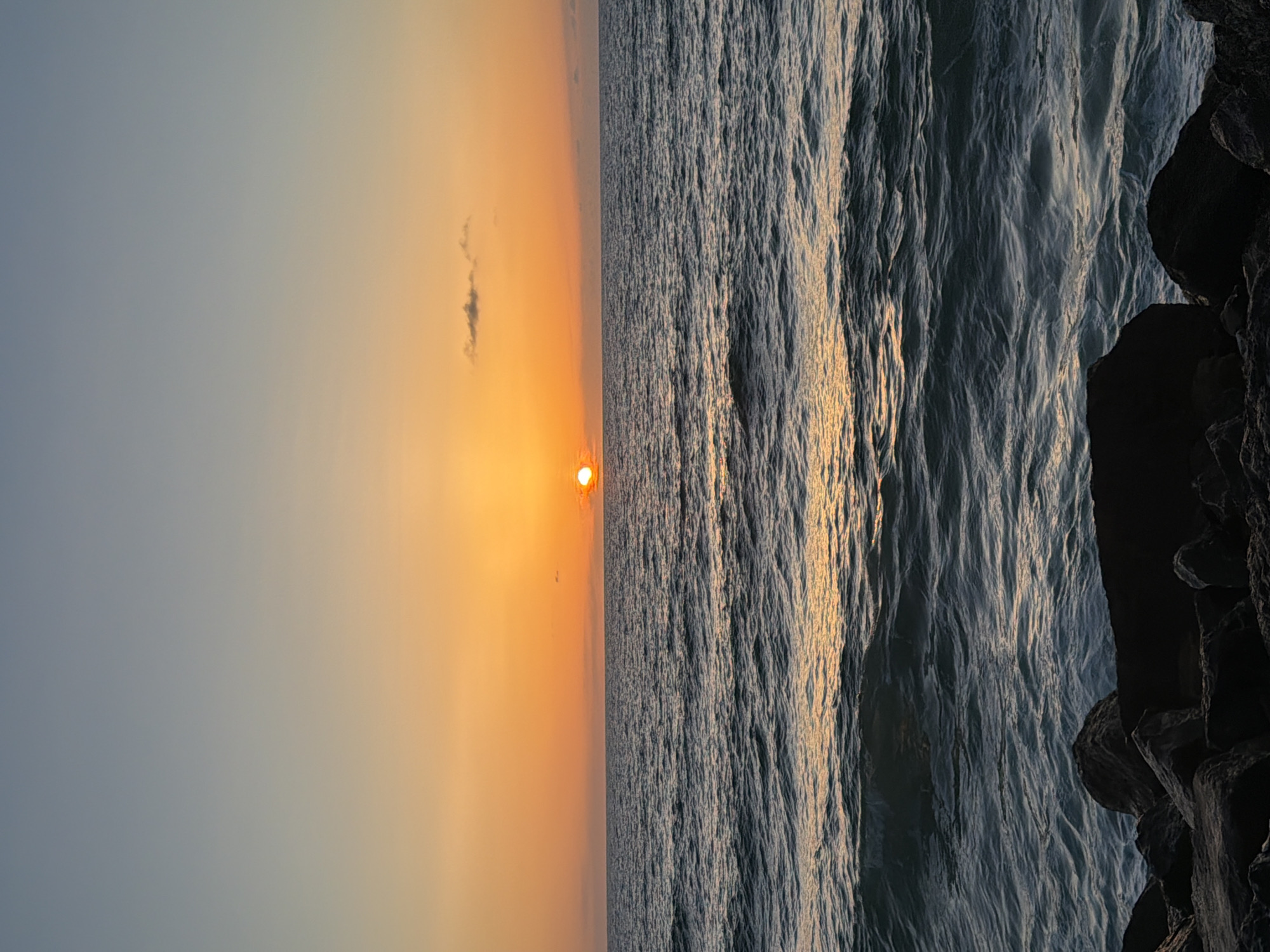The image depicts a sunset over the Mediterranean Sea, likely taken in Tel Aviv, Israel. The sky is a gradient of blue to orange, with the sun visible near the horizon. The sea is textured with waves, reflecting golden light from the setting sun. In the foreground on the right, there are dark, rocky structures. There are no people visible in the image.
