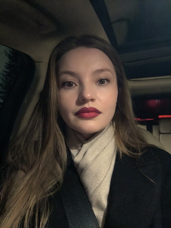 A woman takes a selfie in a car at night. Her light brown hair falls past her shoulders. She wears a black coat over a cream-colored scarf. A black seatbelt crosses her chest. She has fair skin, neutral-toned eyeshadow, and wears a deep red lipstick. The car's interior is visible, including the roof and leather seats. A red light glows in the background, adding to the nighttime ambiance. Outside the car, a dark, blurry shape suggests a tree.