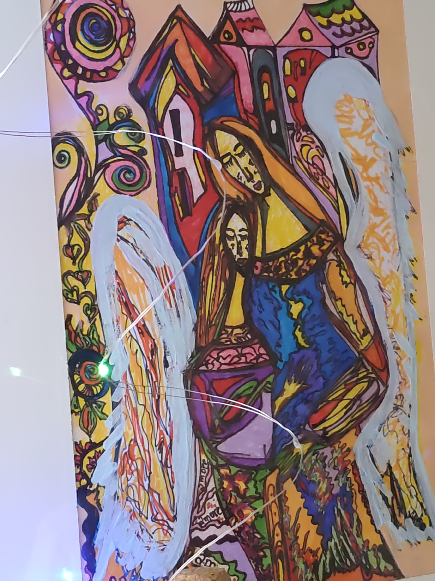 The image displays a colorful, abstract painting featuring two figures that appear to be angels. The figures have large wings and are rendered in bold, expressive lines and vibrant colors, including blues, yellows, oranges, and purples. The background suggests a cityscape with stylized houses. The painting is illuminated by a string of small, multi-colored LED lights draped across its surface. The image quality is somewhat blurry, and the focus is soft, with a slight tilt to the right. The painting is likely a piece of folk art or art nouveau. There is no discernible location within Rostov-on-Don, Russia, or any specific context provided by the image itself.
