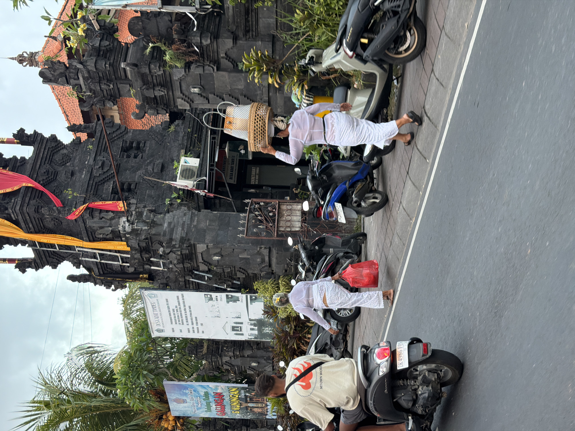 This is a street scene in North Kuta, Indonesia, capturing a moment in front of a traditional Balinese gate. On the left, a man rides a scooter, facing away from the camera. To his right, a woman in traditional white attire walks, carrying a red bag in her right hand. Further right, another woman, also in white, walks away from the camera, balancing a large woven basket filled with items on her head. Behind them, a series of parked scooters line the sidewalk, in front of a dark stone gate adorned with carvings. Yellow and red fabric banners hang from the gate structure. A large white sign with Indonesian text is posted to the left of the gate. Palm trees and other lush vegetation are visible behind the gate. The sky is overcast.