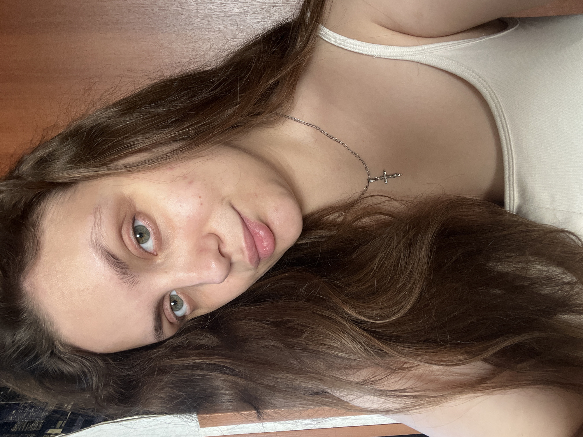 A young woman with long brown hair and green eyes is lying down, wearing a white tank top and a silver necklace with a cross pendant. She is looking at the camera with a slight smile. The background appears to be a wooden surface and a patterned sheet. The location is unknown, and there is no information in the image to confirm it is in Ольховка, Russia.