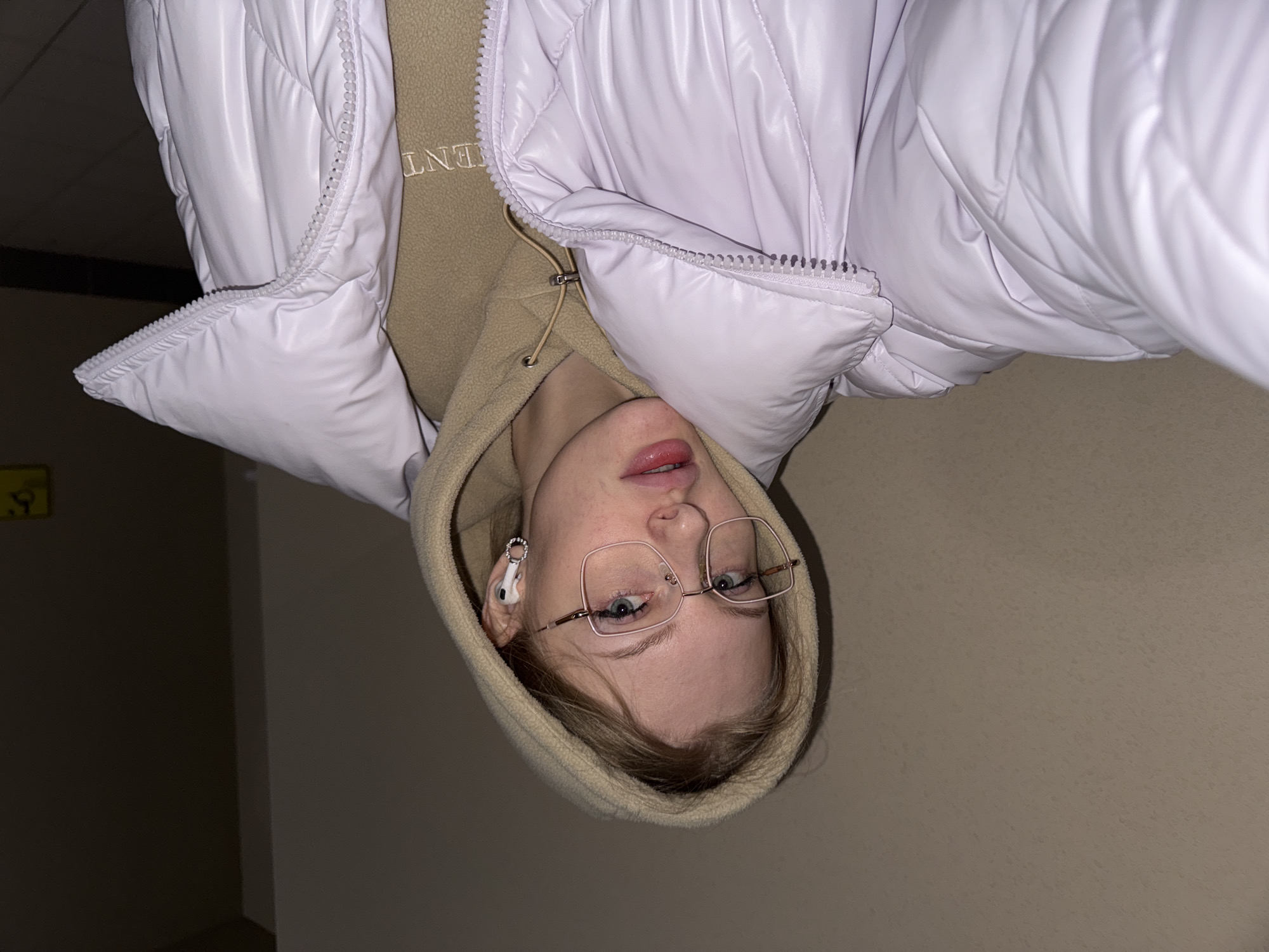 The image is a close-up portrait of a young woman, initially presented upside down, featuring her from the chest up. She has light-colored hair, light blue-grey eyes, and is wearing clear-framed glasses. Her mouth is slightly open, revealing a hint of teeth, and her lips are a glossy pink. A white earbud is visible in her left ear.
She is dressed in a shiny, light lavender or pale purple puffer jacket, which is unzipped. Beneath the jacket, she wears a beige hooded sweatshirt, with the hood pulled up around her head. Part of a word, "ENT," is visible embroidered or printed on the front of her sweatshirt.
The background consists of plain, muted beige or light brown walls, indicating an indoor setting, possibly a hallway or room. There is a darker corner visible on the left side of the frame with a faint, blurry yellow object or sign. The lighting appears even, suggesting indoor artificial light. The woman is looking directly at the camera with a neutral to slightly inquisitive expression.