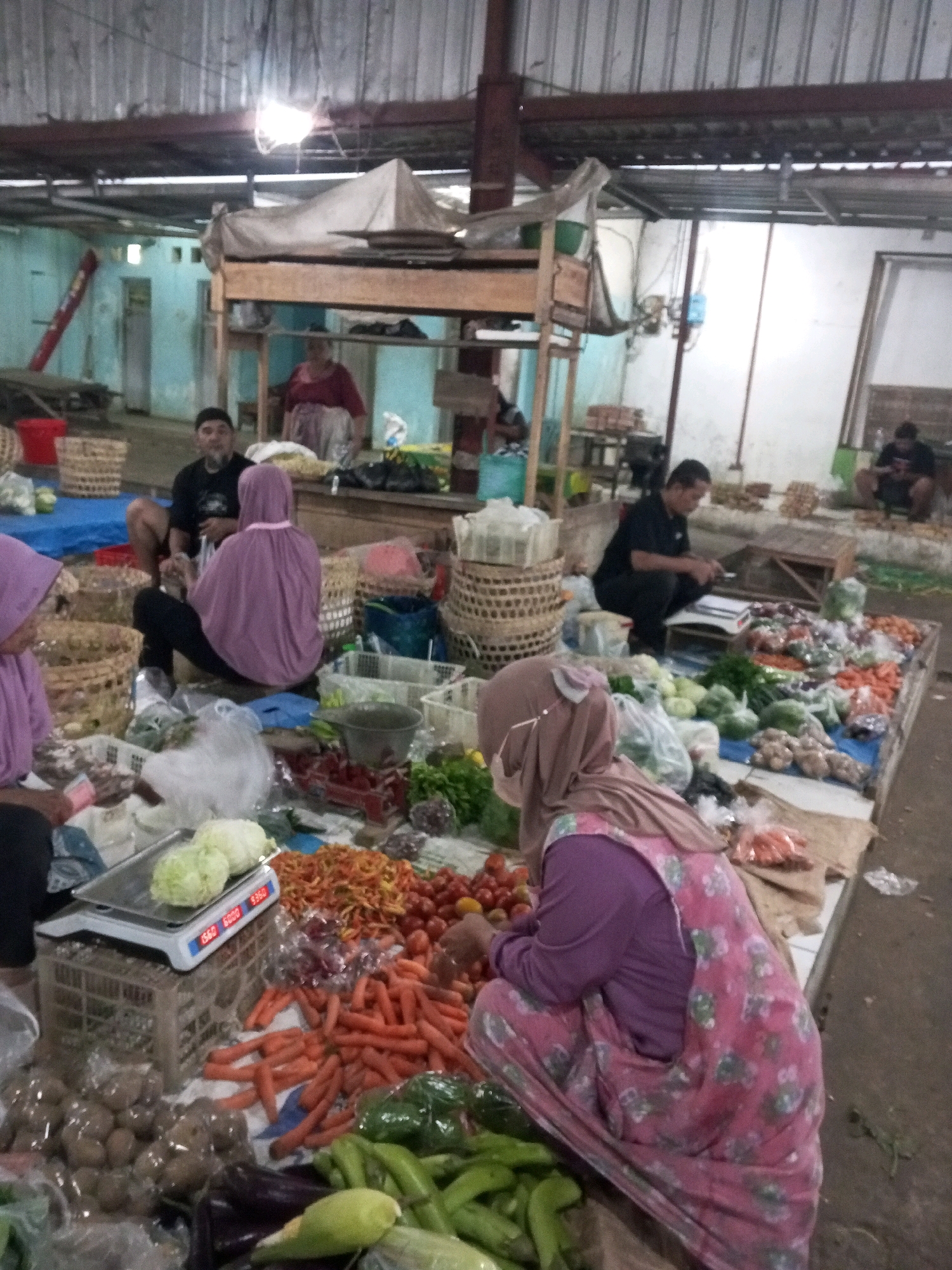 The image depicts a busy marketplace scene in Gentan, Indonesia, likely an outdoor or semi-covered area with a corrugated metal roof. In the foreground, a woman wearing a light purple hijab and a pink floral apron is crouching down, arranging produce on a flat surface. She appears to be selling vegetables and fruits, with various items like cabbages, carrots, tomatoes, and potatoes displayed on tables and in baskets. A digital scale sits near the cabbages, indicating the presence of a transaction. To the left, another woman in a purple hijab is also seated, holding what appears to be money. In the mid-ground, several individuals are present. A man wearing a black t-shirt and a black cap is seated, looking down at something in his hands. Another man in a black t-shirt and dark pants is crouching, seemingly engaged in writing or sorting documents on a table. Behind them, more stalls and produce are visible, along with woven baskets and tarps. The overall environment suggests a vibrant local market with people actively participating in commerce.
