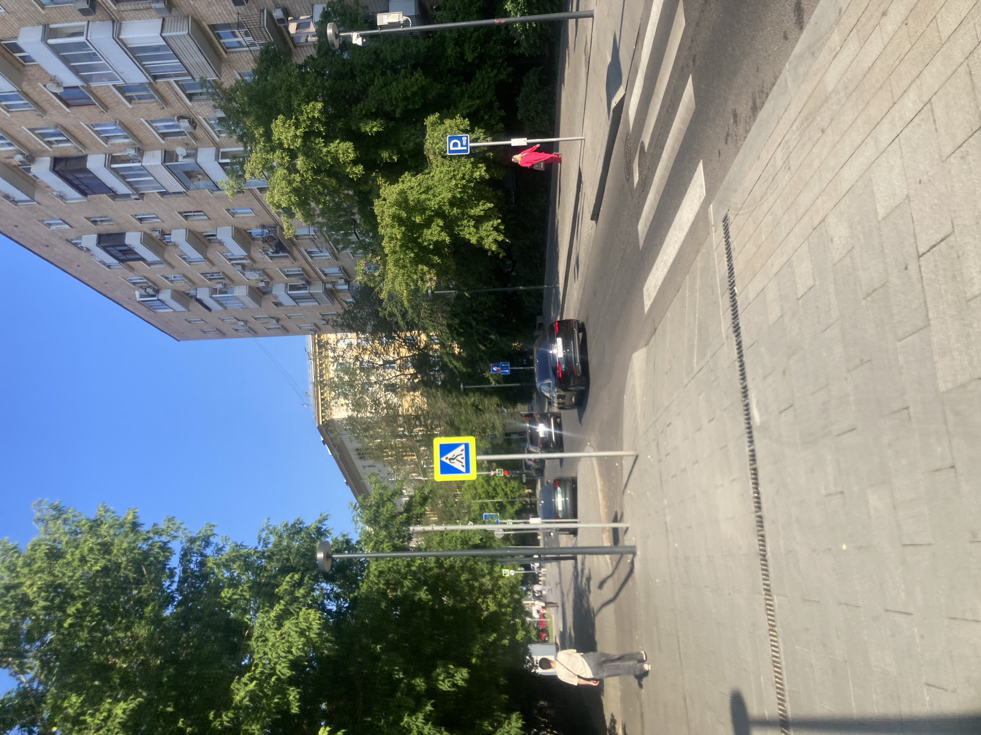 A street scene in Moscow, Russia, on a bright, sunny day. The image is dominated by a multi-story brick apartment building on the right, with numerous balconies and windows. In the foreground, a paved sidewalk runs parallel to a street where cars are visible, including a black sedan and a dark SUV. A pedestrian crossing with white stripes is marked on the road.
On the sidewalk, a person wearing a light-colored t-shirt and gray pants walks away from the camera. Further down the sidewalk, a person in a bright red dress is walking towards the camera. There are several streetlights and pedestrian crossing signs visible. Green trees line the street, casting shadows on the pavement. The sky is a clear, vibrant blue. The overall impression is of an ordinary urban environment on a pleasant day.