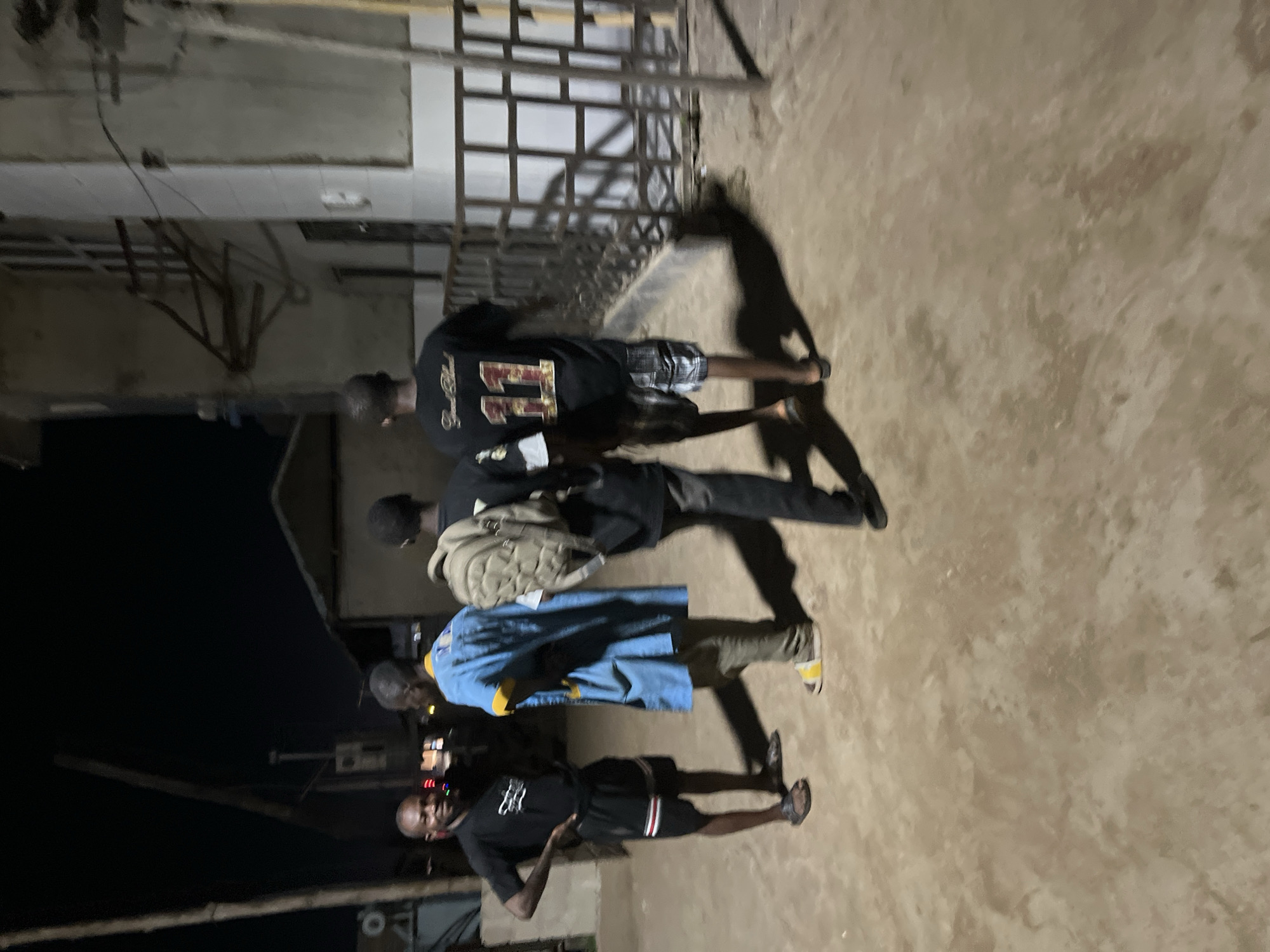The image captures four individuals, appearing to be men, outdoors at what seems to be nighttime. They are standing on a light-colored, rough ground surface, possibly concrete or dirt, with long, distinct shadows cast behind them by an unseen light source, indicative of artificial illumination in the dark.
From left to right in the upright orientation, the first person, in the foreground, wears a black t-shirt with an unclear white text or design on the front and black shorts with white and red stripes. This individual is looking towards the camera with a neutral expression, with one hand on their hip. The second person wears a blue denim-like jacket, khaki pants, and yellow-and-white slide sandals. The third individual is dressed in a dark long-sleeved shirt and dark pants, and carries a light-colored backpack. The fourth person wears a black t-shirt with a large number "11" in a reddish-gold color on the back, paired with plaid shorts.
In the background, there's a building with light-colored walls and a metal grate or railing structure, suggesting an urban or semi-urban setting. The overall atmosphere is dim, illuminated primarily by the artificial light that creates the strong shadows. No specific location cues are clearly visible beyond the general architectural style and ground surface. The individuals appear to be casually gathered or walking together.