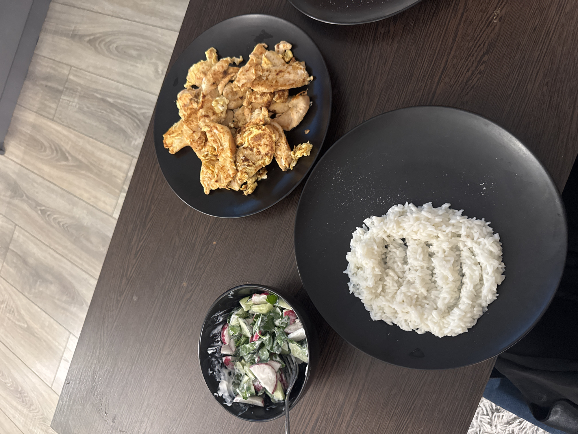 The image depicts a meal consisting of stir-fried chicken, white rice, and a salad. The chicken is presented on a black plate, appearing browned with egg, likely seasoned. The rice is also served on a black plate, positioned on the right side of the frame. A salad, consisting of sliced radishes, cucumbers, and greens in a creamy dressing, is placed in a black bowl on the left side. A fork is visible inside the bowl, suggesting that the salad has been partially consumed. The meal is set on a dark wooden surface. The image does not contain any people. There is no indication of the city of Unknown, Russia.