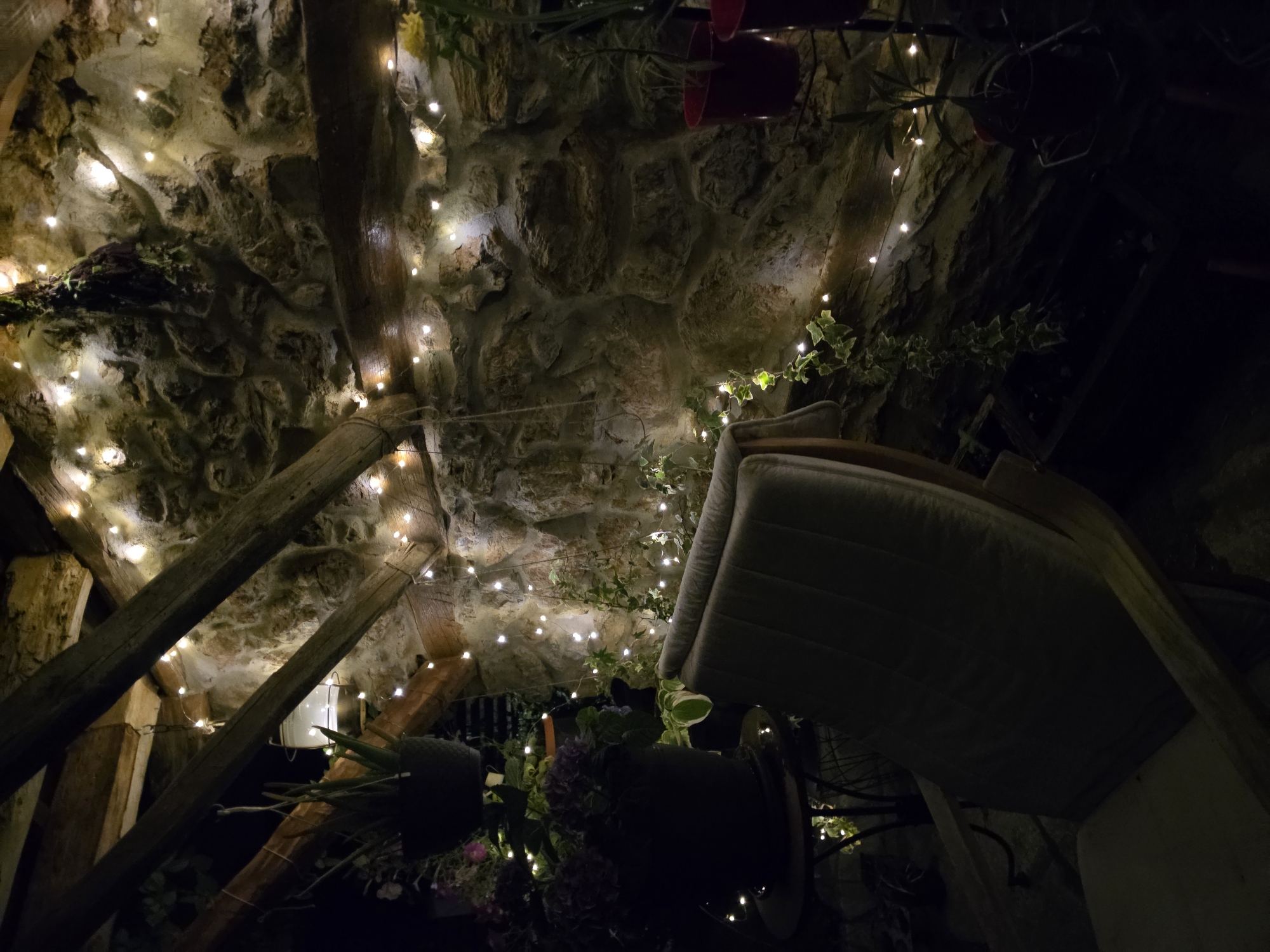 The image depicts an outdoor seating area at night, illuminated by strings of fairy lights. A wooden chair with a padded cushion is positioned in the foreground, facing the viewer. Behind the chair is a rustic stone wall covered in ivy and adorned with more fairy lights. Wooden beams are visible overhead, supporting the roof structure. To the left, potted plants, including a large dark pot with purple flowers and a hanging basket with a succulent, are placed on a small table. The overall atmosphere is cozy and inviting, suggesting a tranquil evening setting. The location is not specified beyond "Unknown, Unknown."