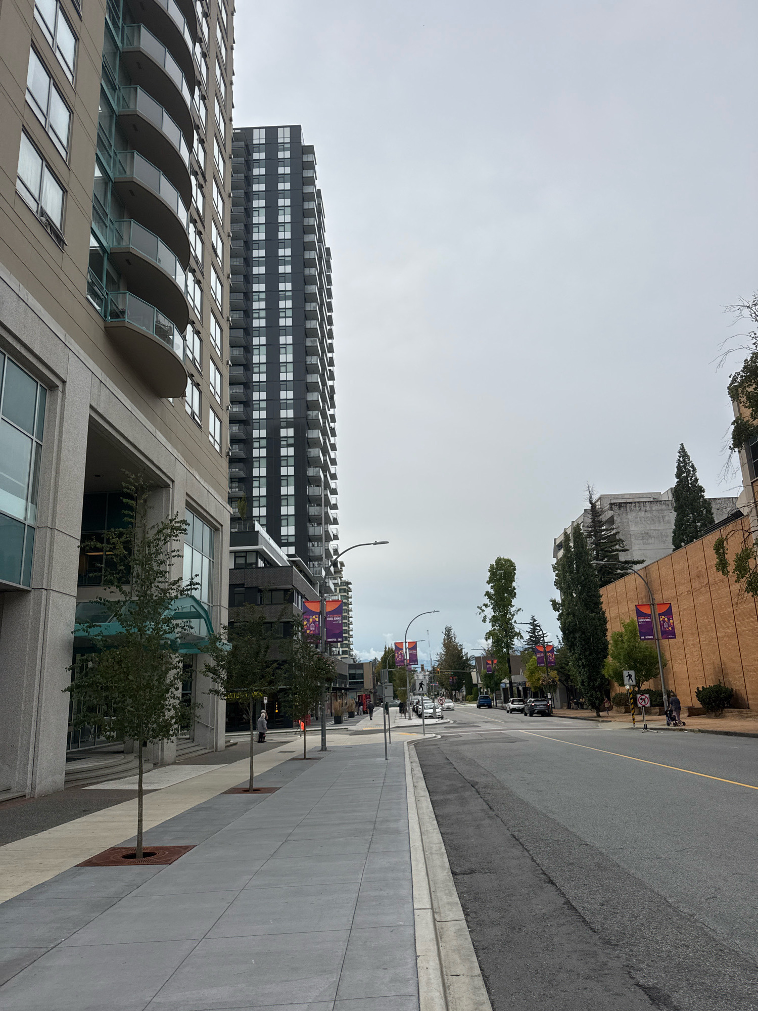The image shows a street scene in New Westminster, Canada. On the left is a modern beige building with curved balconies. To the right of it, a tall, dark-colored building with many windows and balconies is visible. A wide sidewalk with trees in square planters runs along the buildings. The street itself is asphalt with a yellow line marking the center. There are several cars driving on the street, and a few people walking on the sidewalk. In the background, more buildings and trees are visible under a cloudy sky. Purple and orange banners hang from lampposts along the street. The general impression is a commercial or mixed-use urban environment.