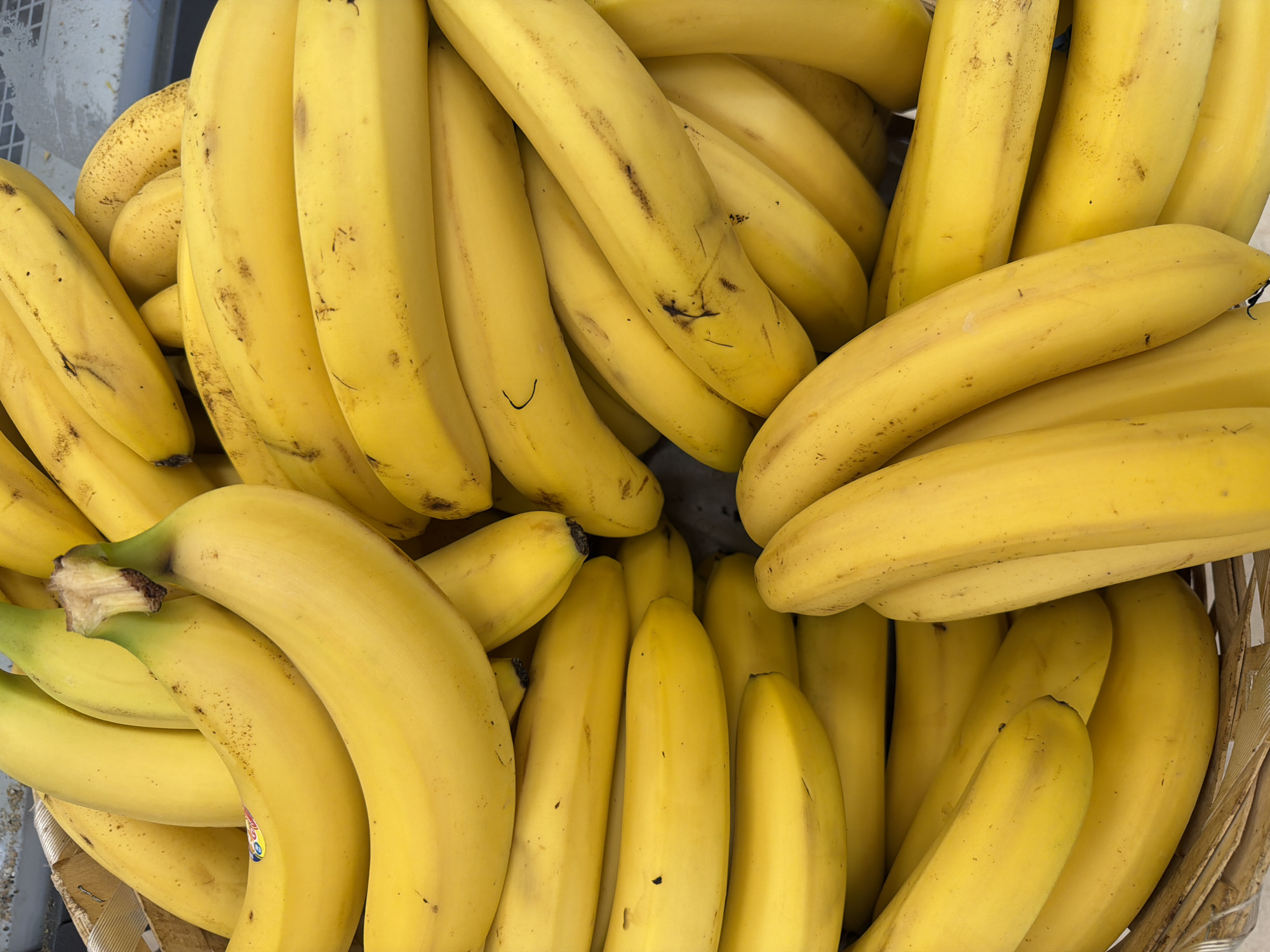 A close-up, overhead shot captures a tightly packed collection of ripe bananas in a woven basket, likely at a market in Kemer, Turkey. The bananas are a vibrant yellow, with some showing minor brown spots, indicating their ripeness. The perspective emphasizes the curved shapes and smooth textures of the fruit, with some stems and a portion of the basket's edge visible. The overall impression is one of abundance and freshness.