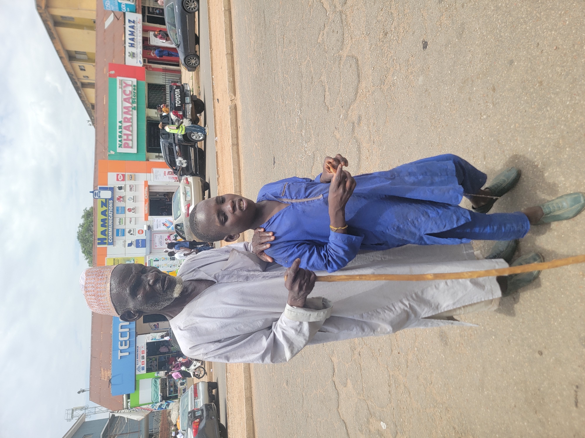 An eye-level medium shot captures an elderly man with a walking stick standing with a young boy wearing a blue robe on a paved sidewalk or street. The man is fair-skinned with a brown and white patterned hat and light grey robe. The boy is dark-skinned with a blue robe and green shoes. The background shows a busy street scene in Jos, Nigeria, with stores like "NASARA PHARMACY & Stores" and "HAMAZ COMMUNICATIONS" visible behind them, along with cars and motorcycles. The perspective is slightly tilted, and the day appears sunny.